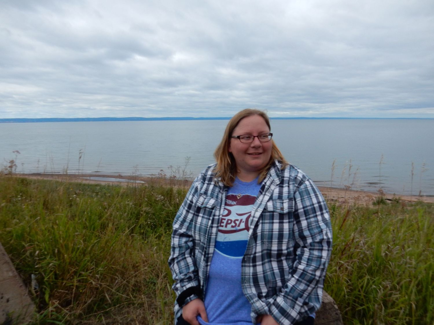 Woman in plaid shirt and Pepsi tee smiles outdoors near grassy shoreline with calm sea and cloudy sky behind her.
