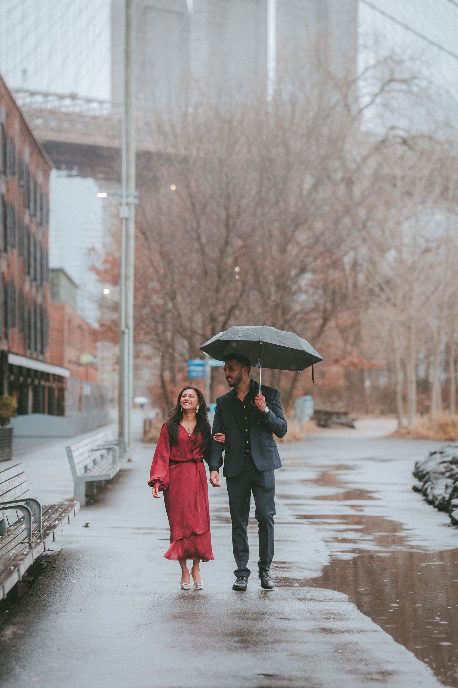 Couple walking under an umbrella with the Brooklyn Bridge in the background on a cold, rainy day in DUMBO, Brooklyn