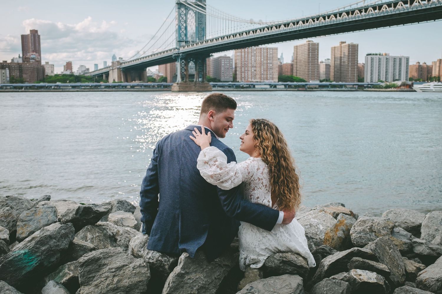 A recently engaged couple embraces while sitting on rocks with a view of the Manhattan Bridge