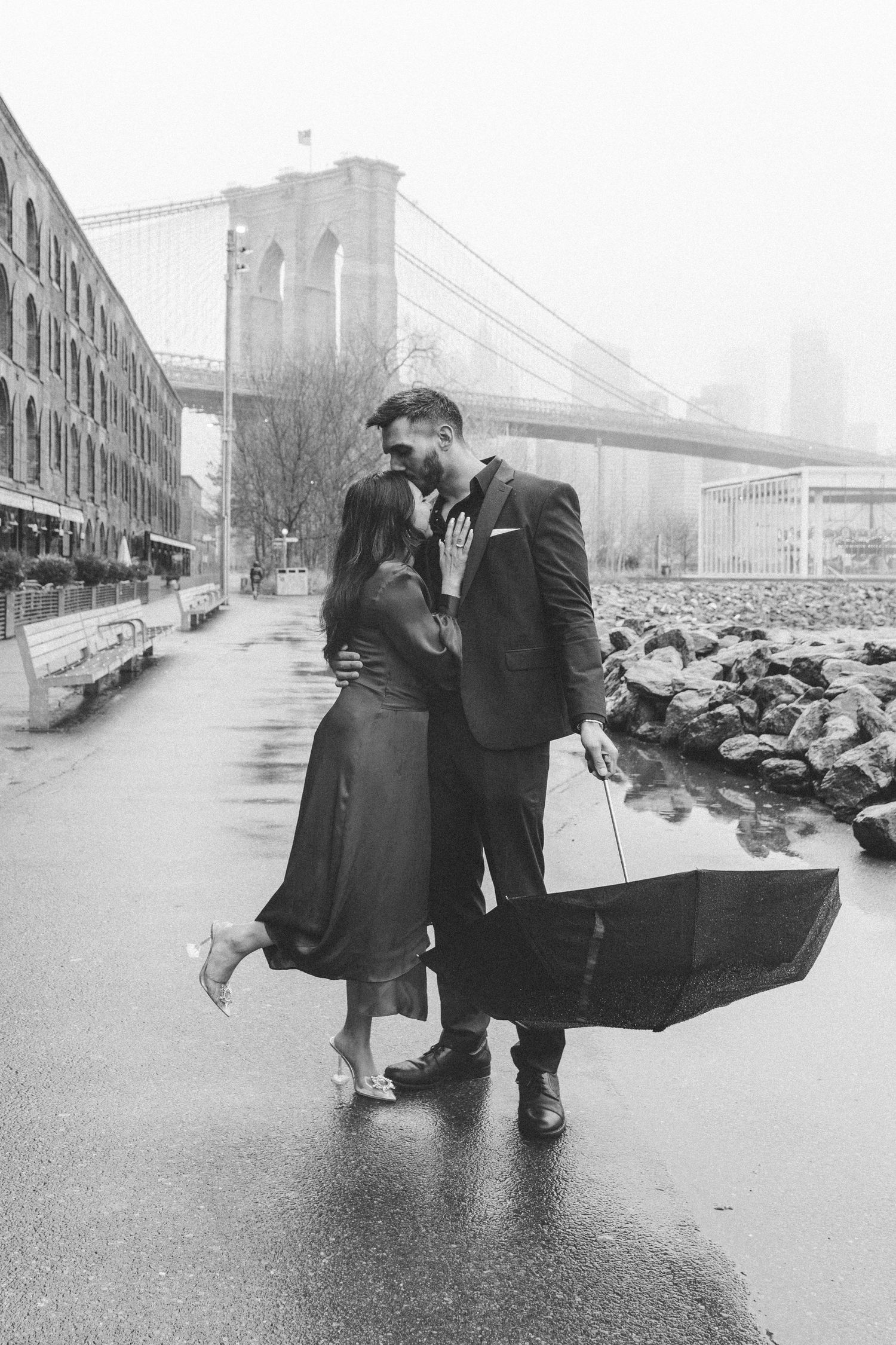 Couple hugging, holding an umbrella with the Brooklyn Bridge in the background on a cold, rainy day in DUMBO, Brooklyn