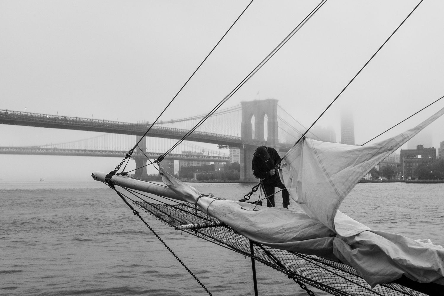 A man working on the sails of a ship docked at South Street Sea Port with the Brooklyn Bridge in the distance