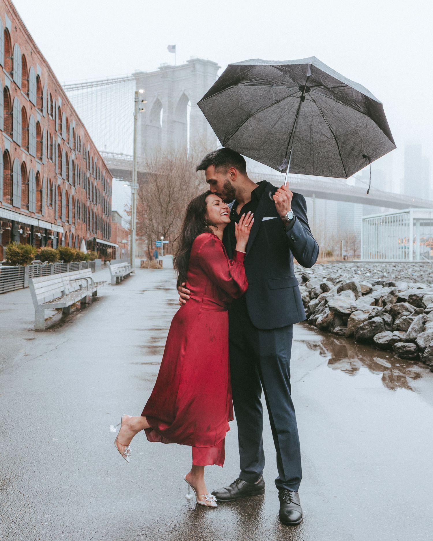 Couple embracing under an umbrella with the Brooklyn Bridge in the background on a cold, rainy day in DUMBO, Brooklyn