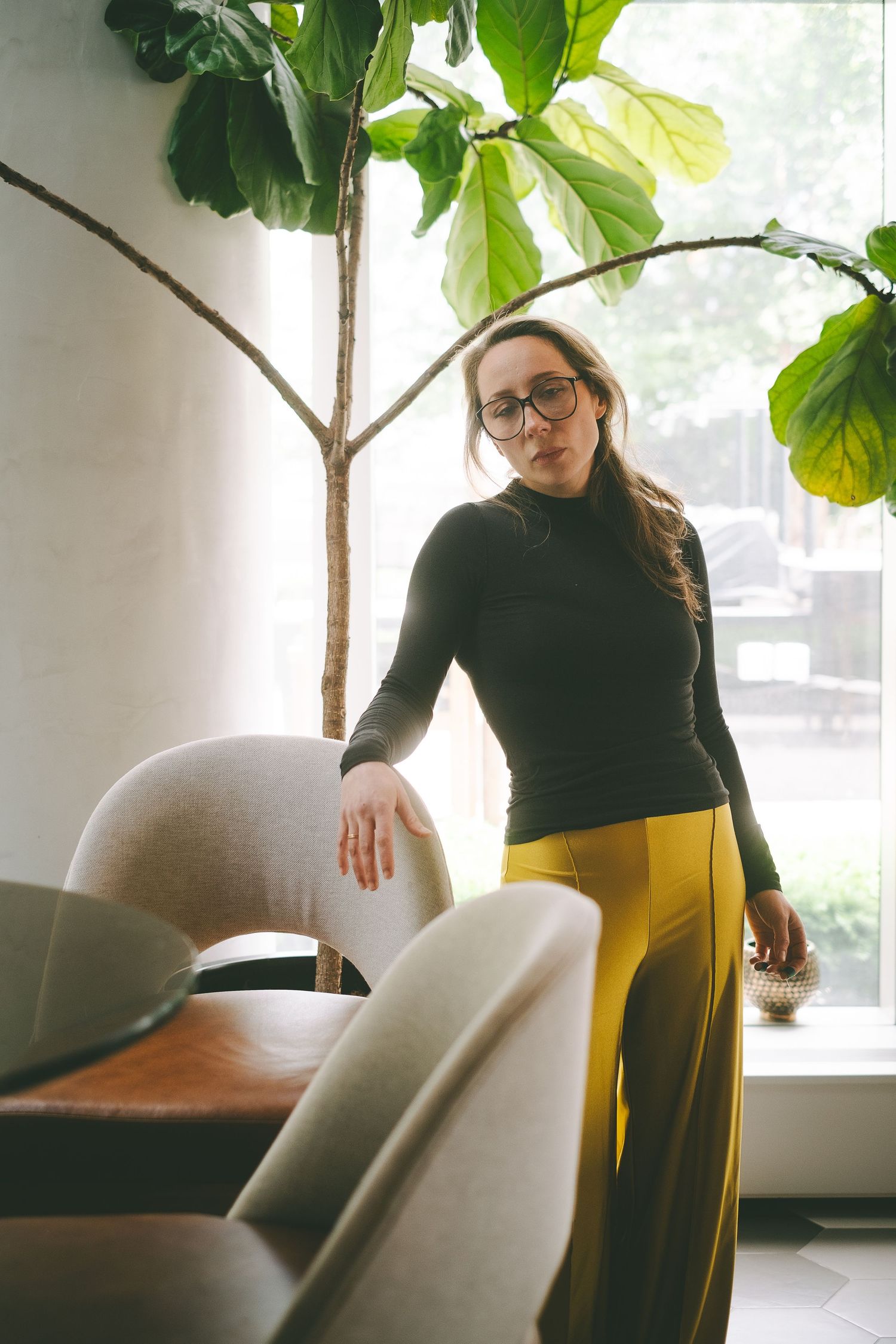 A woman wearing glasses, yellow pants and a black top standing near a plant