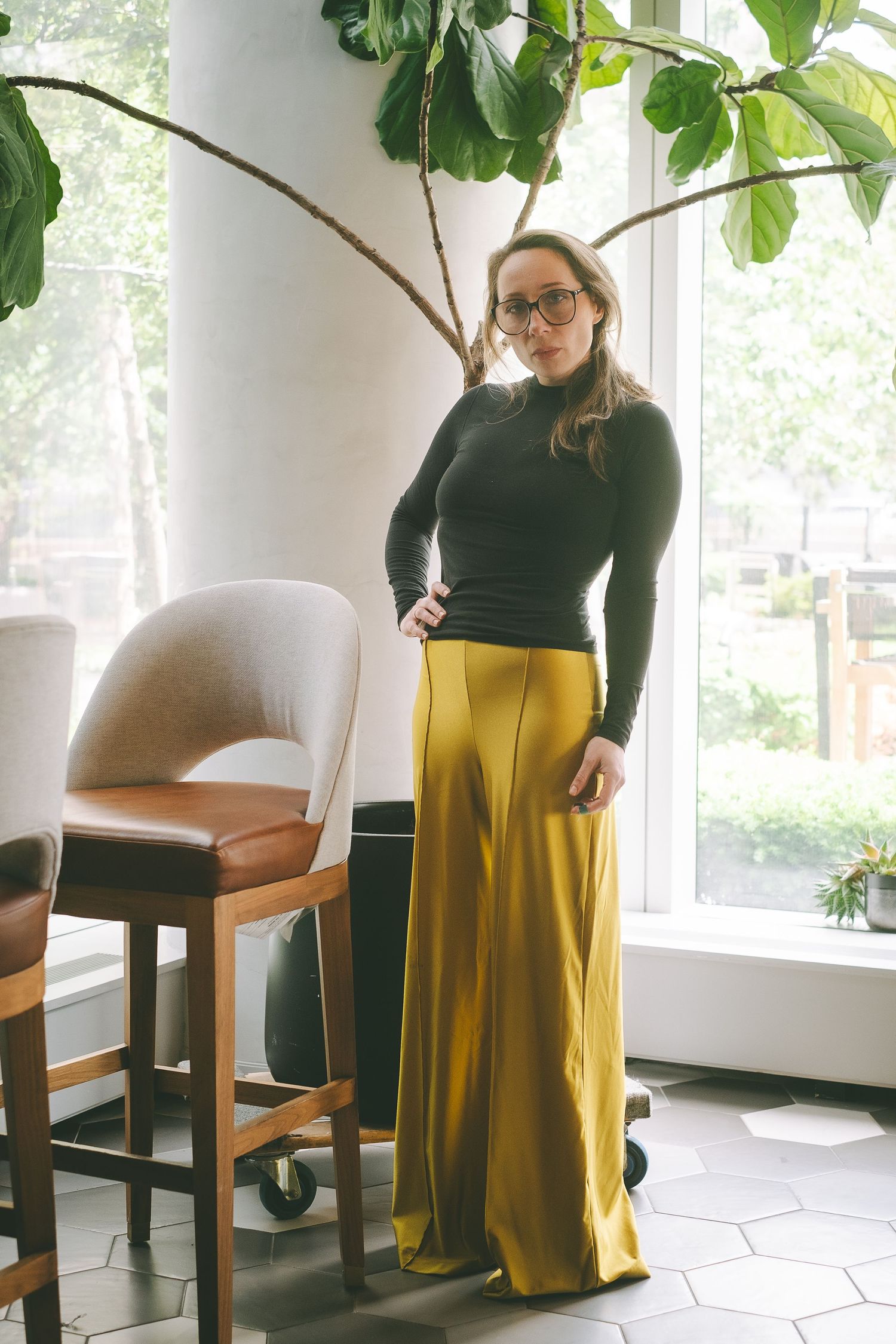 A woman wearing glasses, yellow pants and a black top standing near a plant