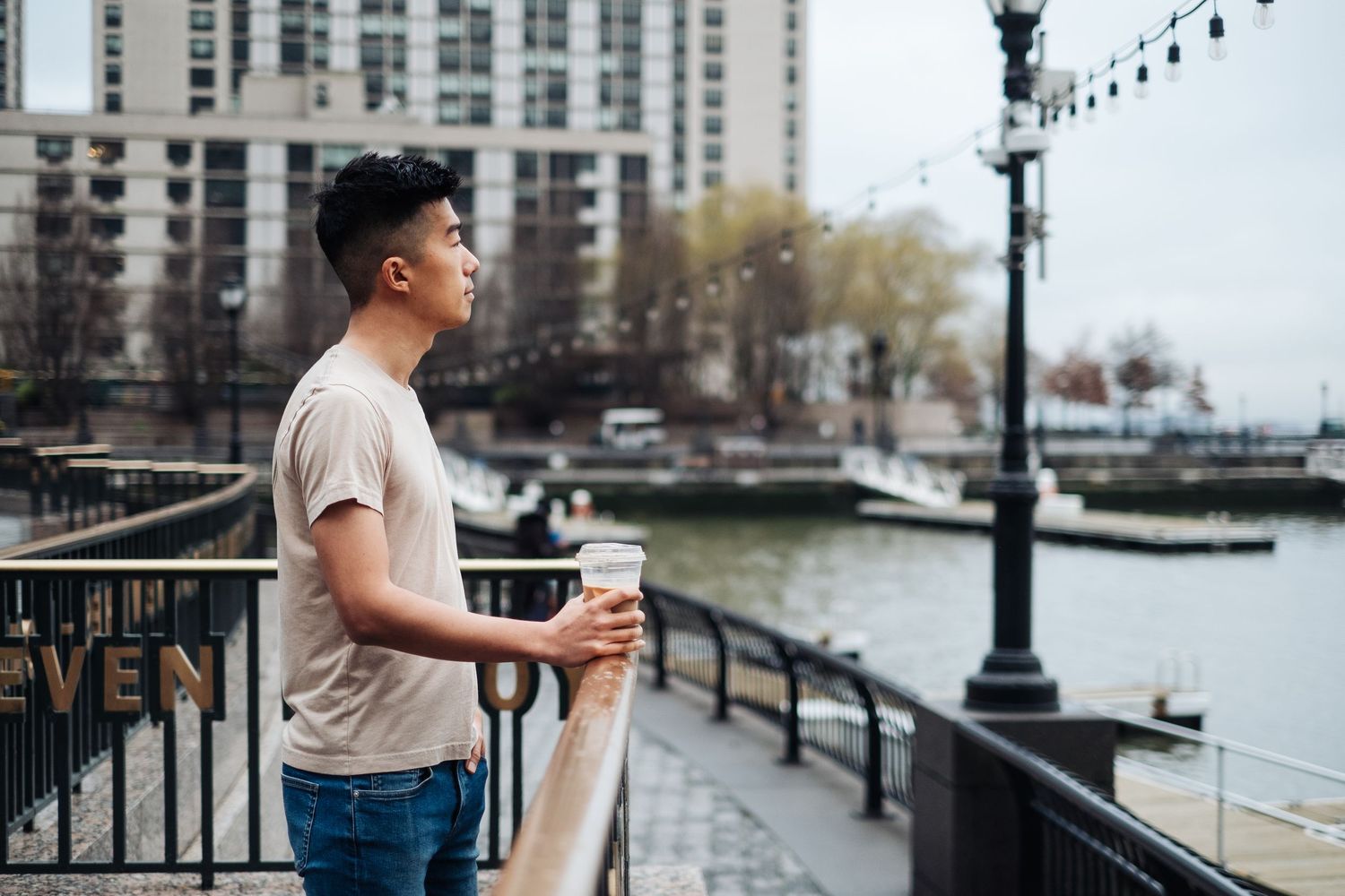 A man in a t-shirt holding a cup of coffee looks out over the Hudson River in New York city