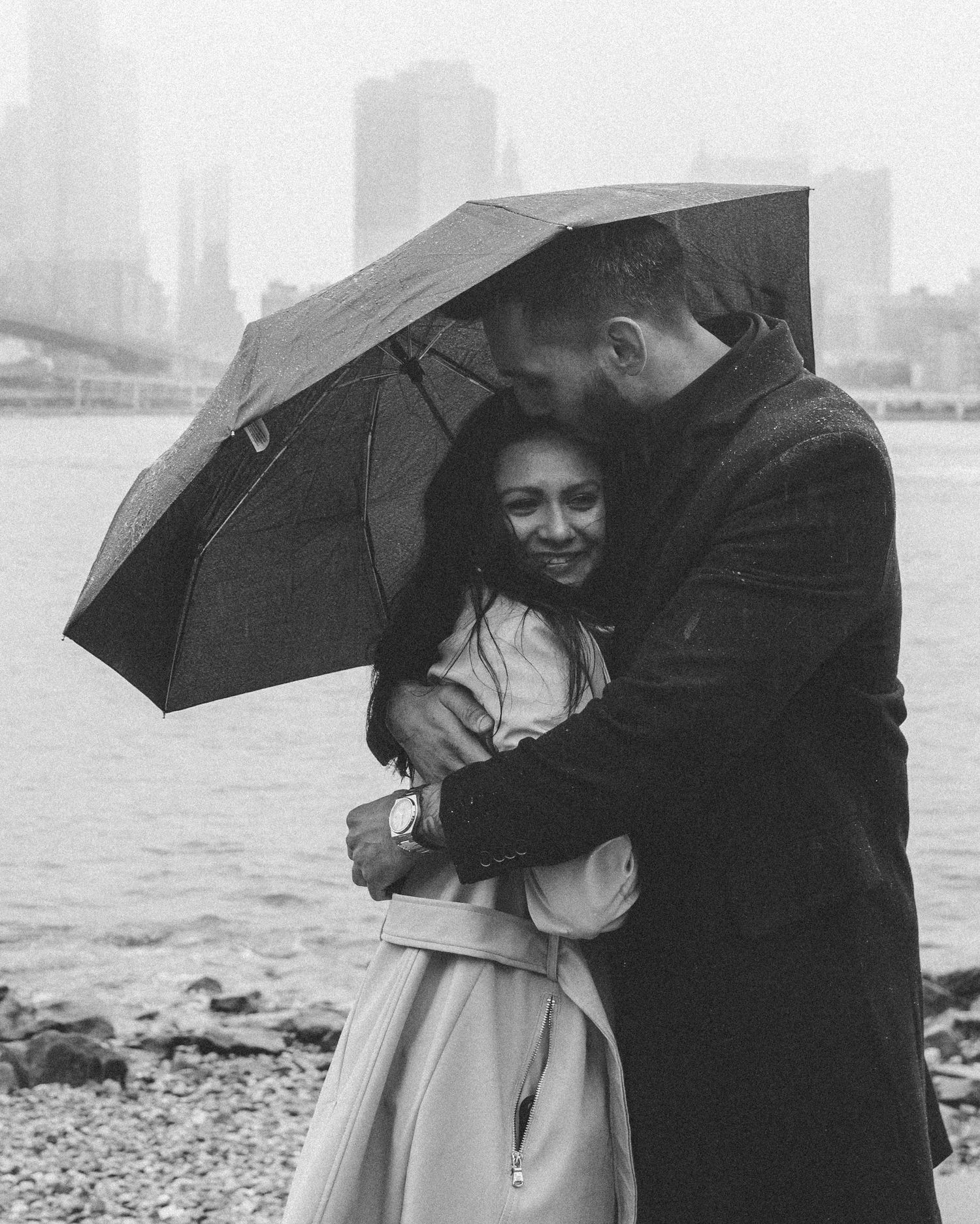 A recently engaged couple embracing under an umbrella on a cold, rainy day in DUMBO, Brooklyn