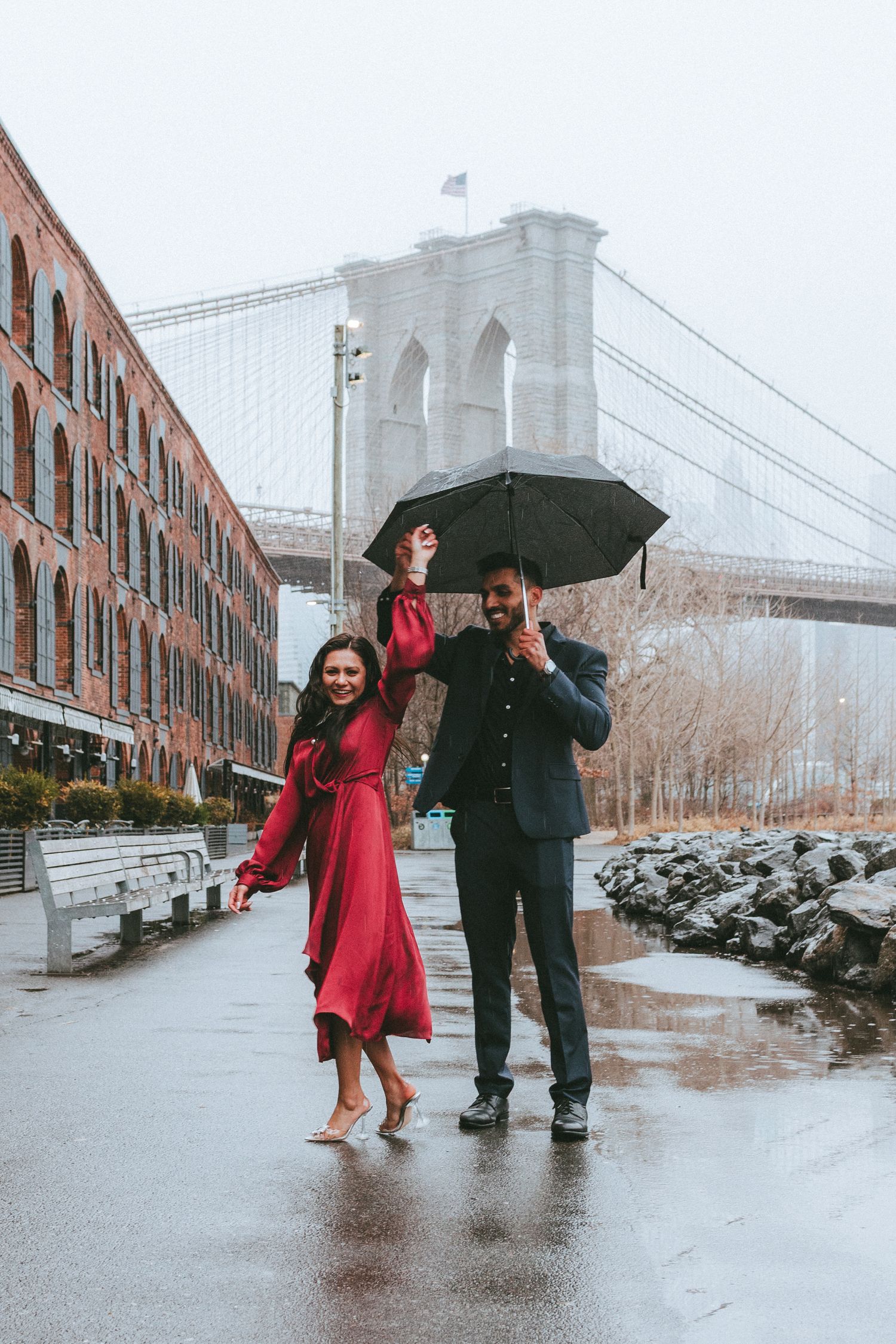 Couple dancing under an umbrella with the Brooklyn Bridge in the background on a cold, rainy day in DUMBO, Brooklyn