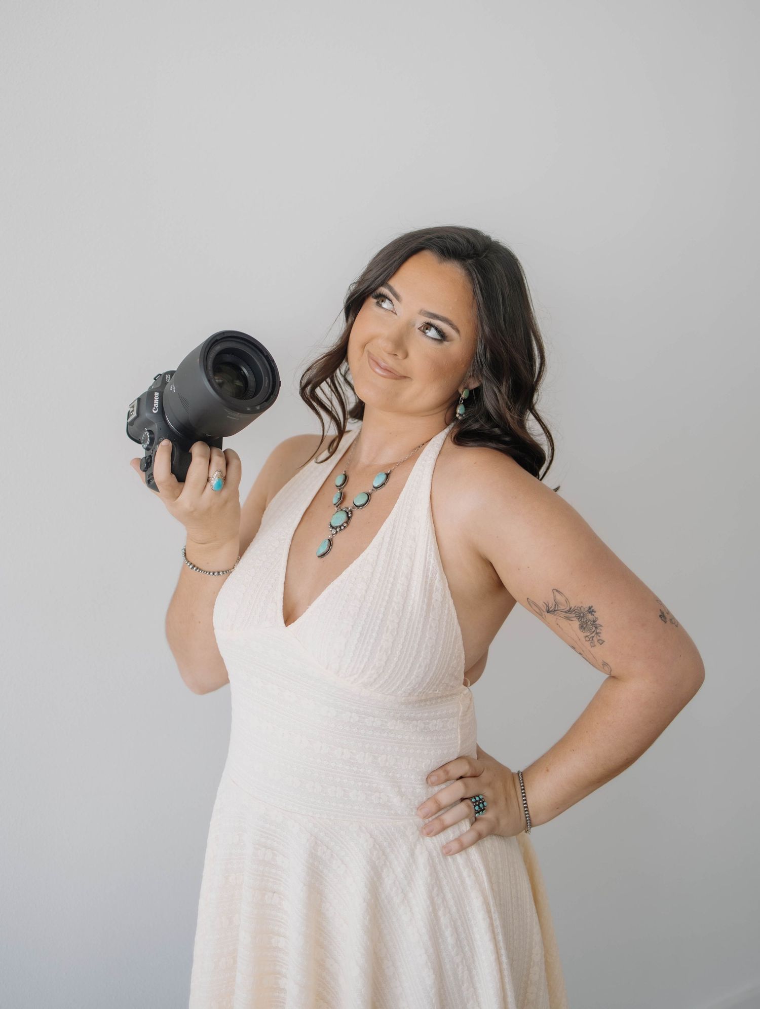 Woman in white dress poses with camera against grey background.