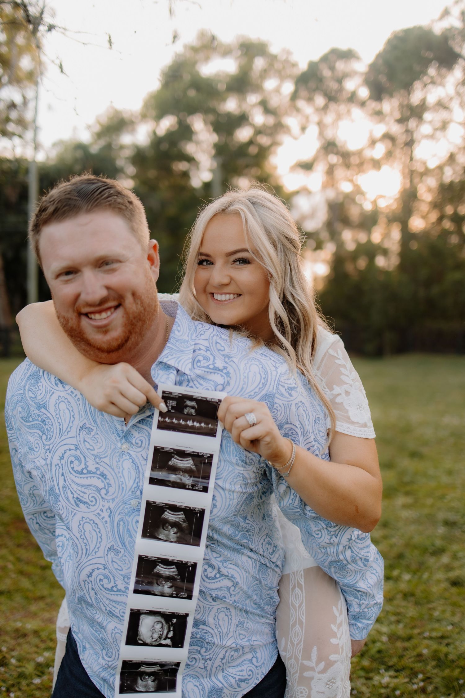 Couple sharing joyful moments while holding ultrasound photos in a sunset field.