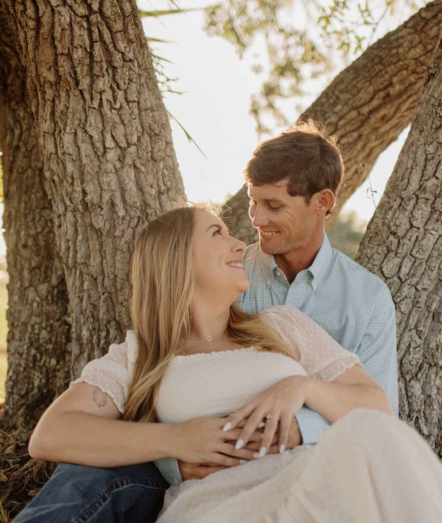 Romantic couple shares tender moments against a sunlit tree backdrop during an outdoor photoshoot.