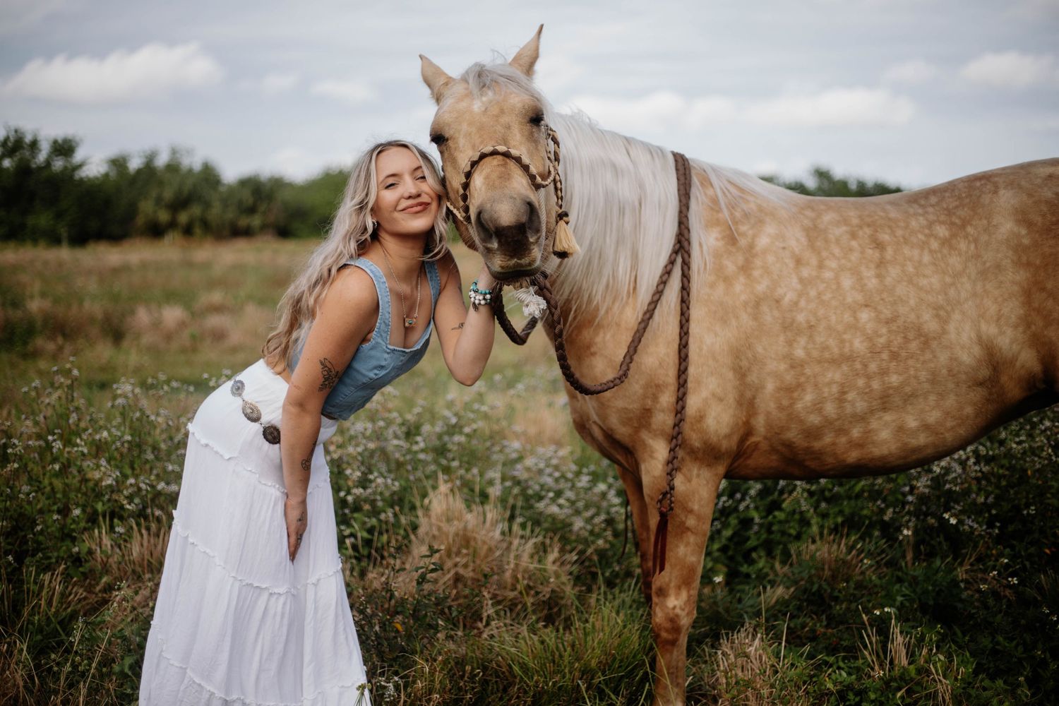 Woman in white dress interacts lovingly with a dappled horse in a rustic outdoor setting.
