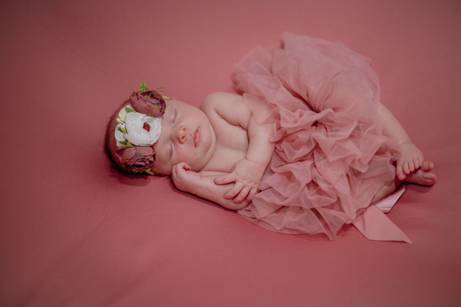 Peaceful sleeping newborn wrapped in pink tulle on rose-colored background.