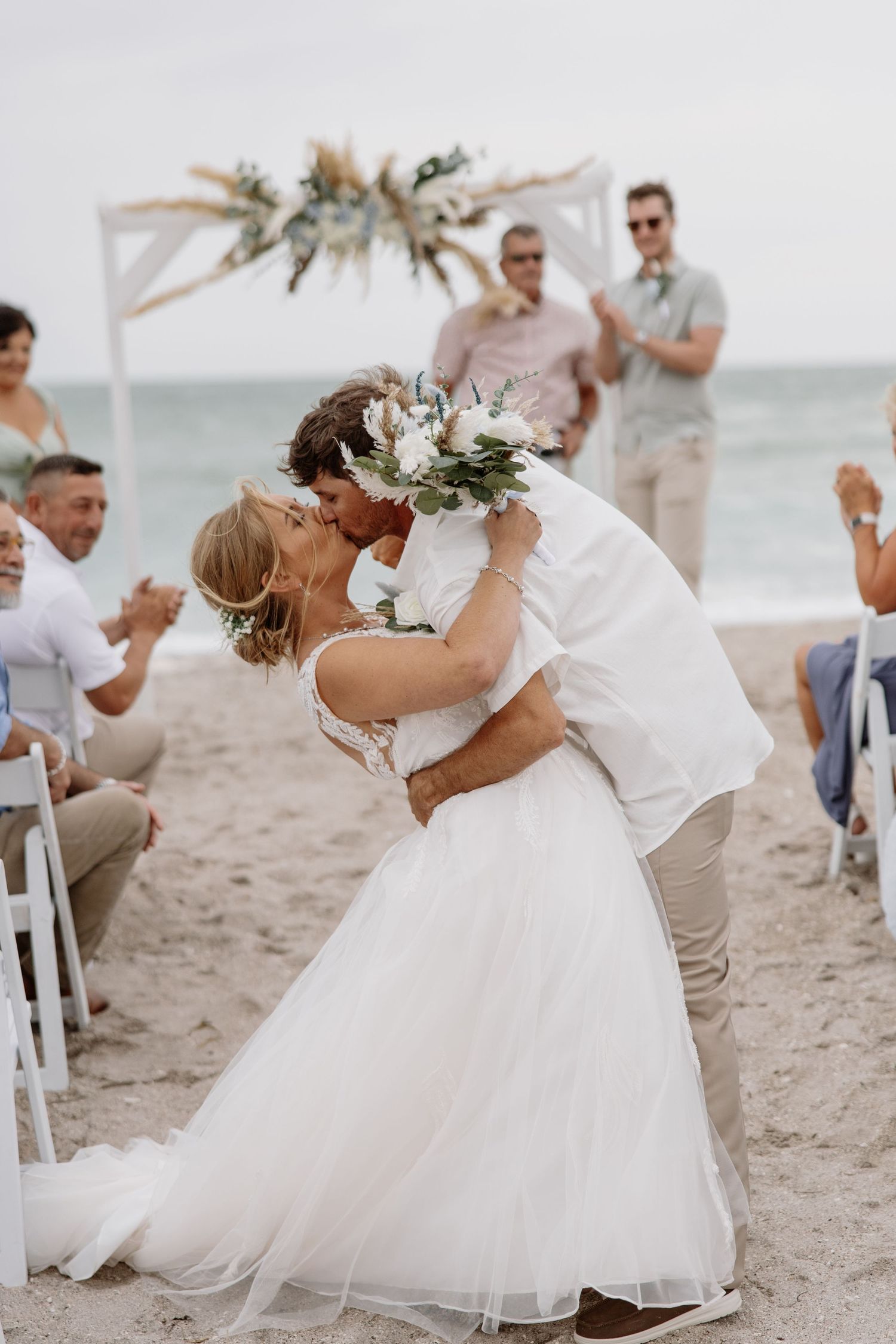 Romantic beach wedding ceremony with white chairs and flowing dresses against ocean backdrop.
