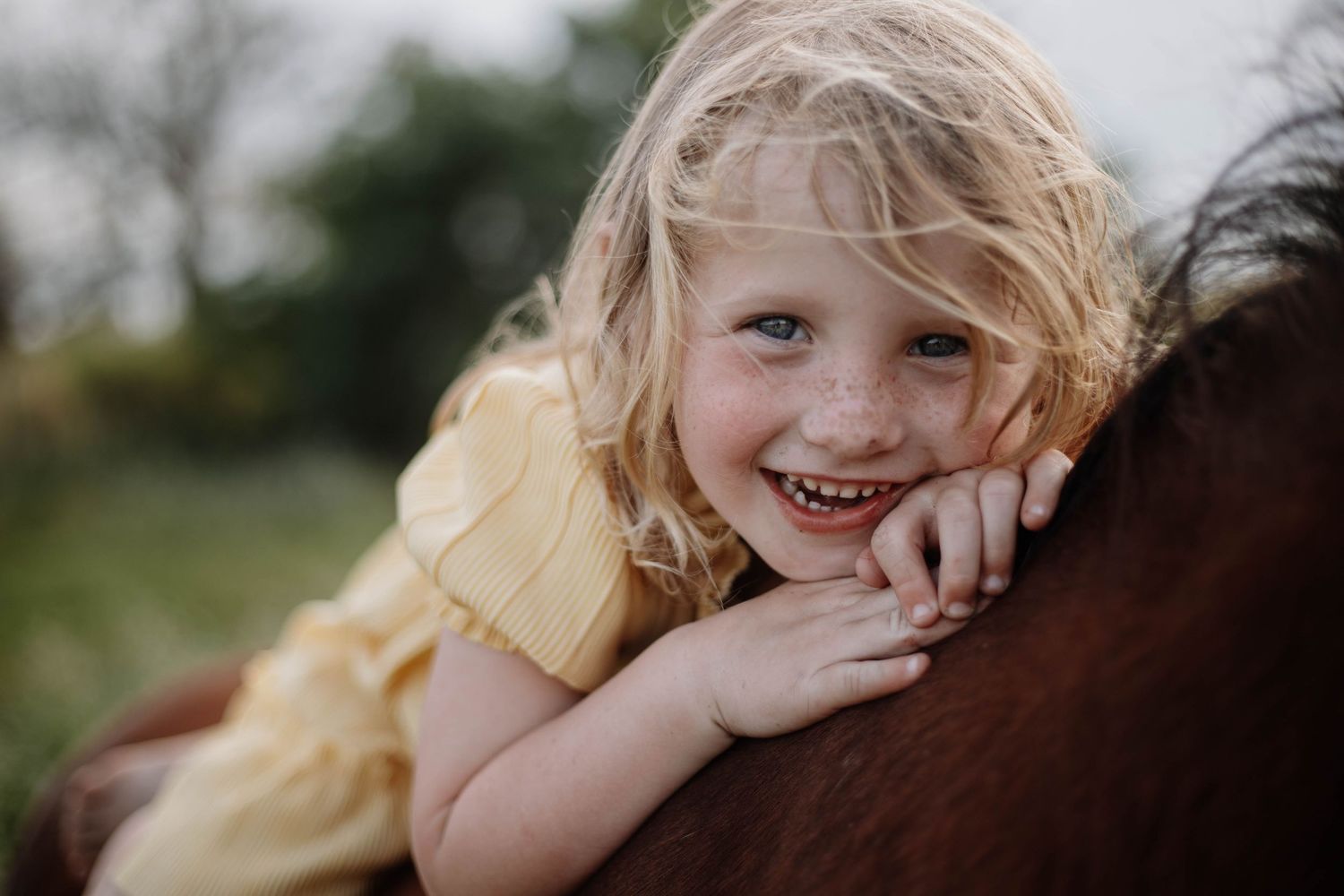 Young child with blonde hair rides horseback while smiling joyfully outdoors.