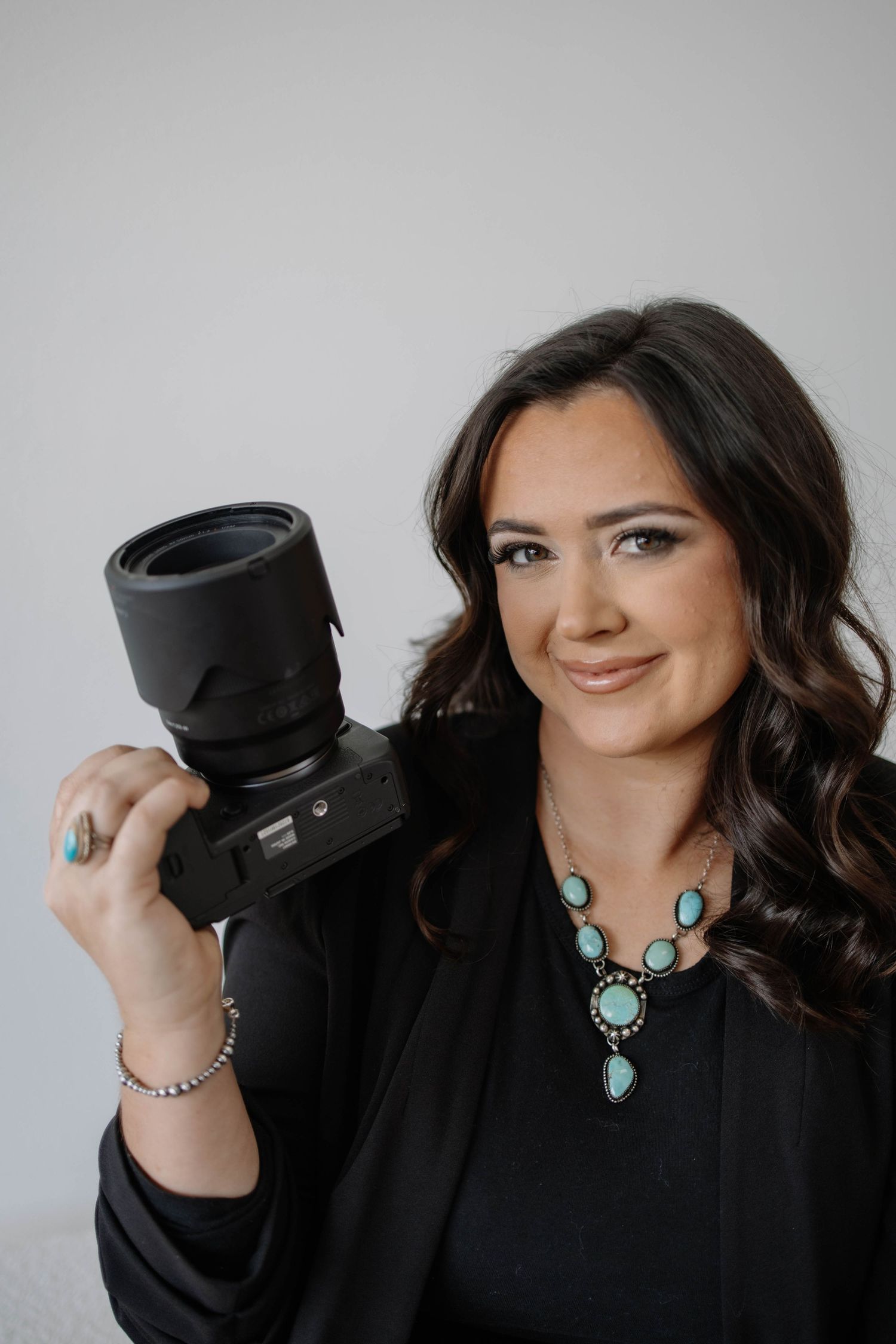 Woman in black top with turquoise necklace poses playfully with camera.