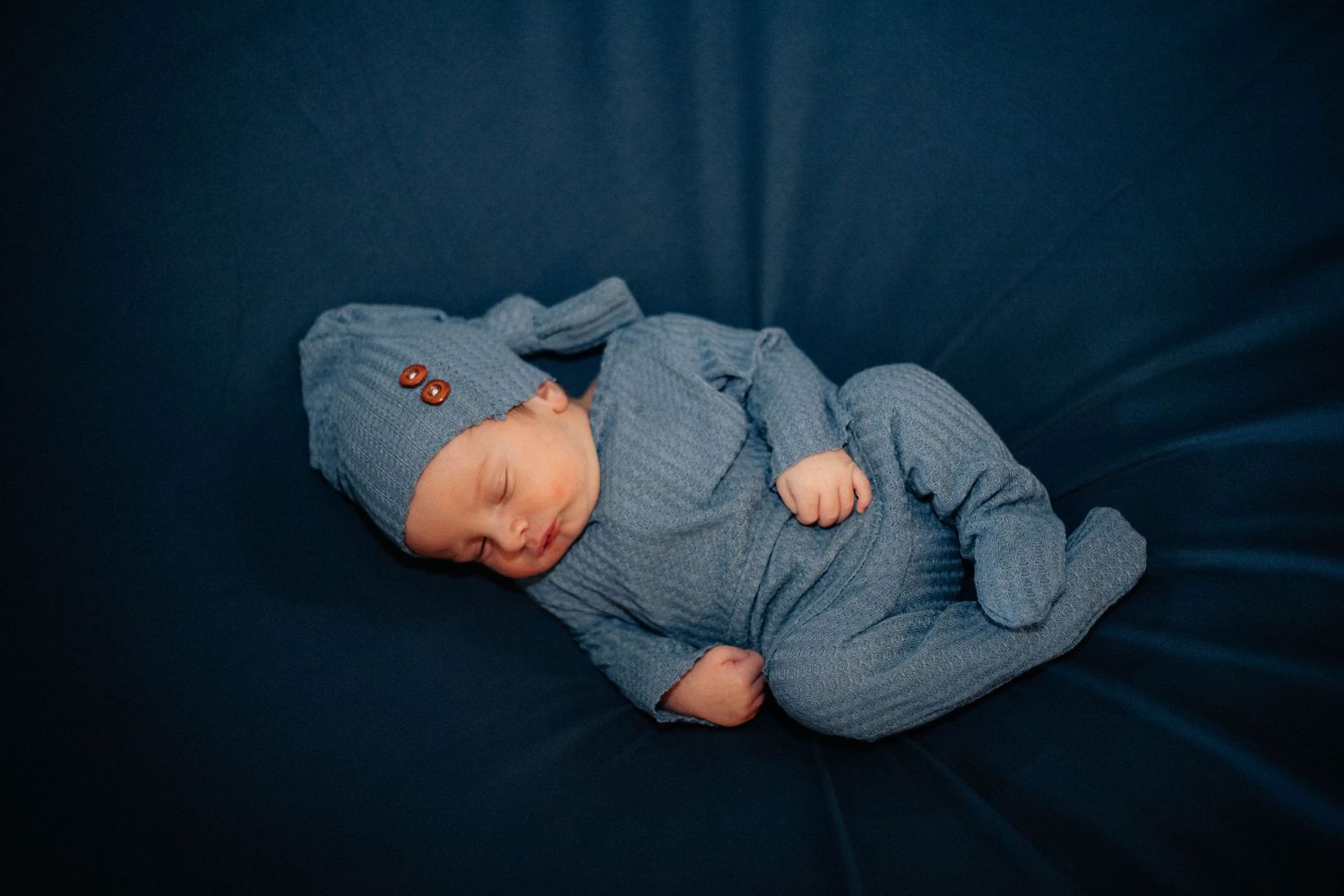 Newborn baby wearing soft blue knit outfit sleeping on dark background.