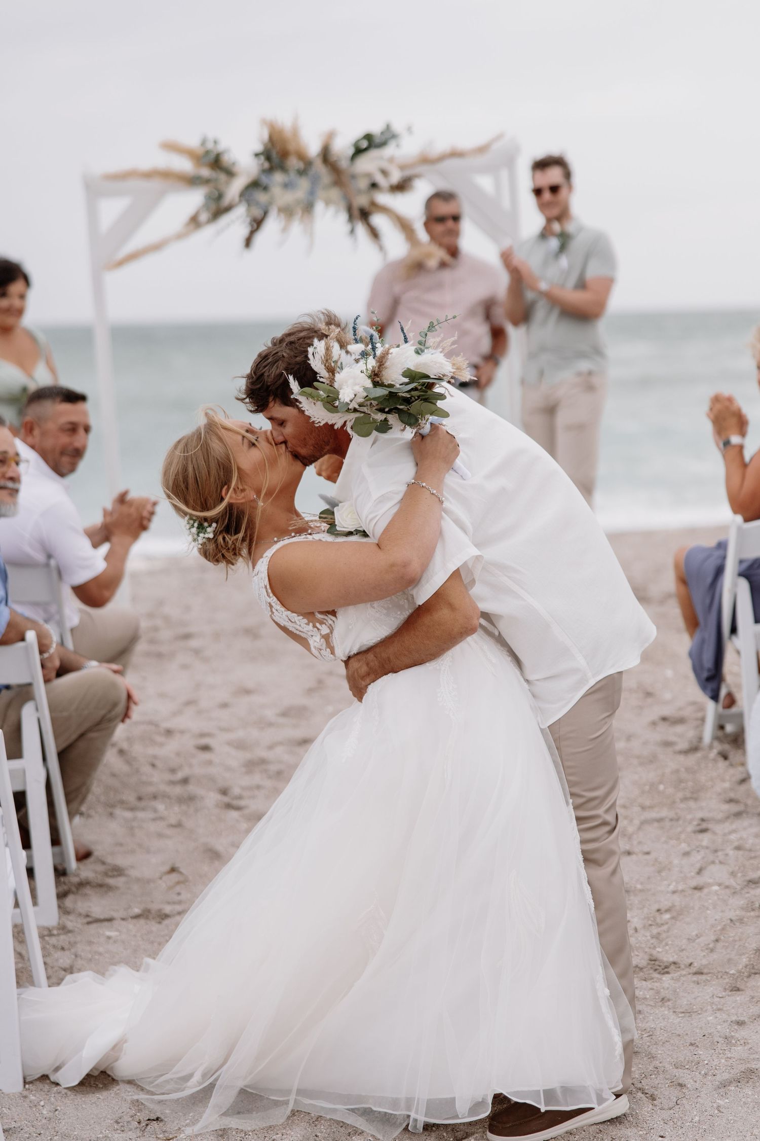 Newlyweds share romantic kiss on beach during sunset ceremony with guests watching.