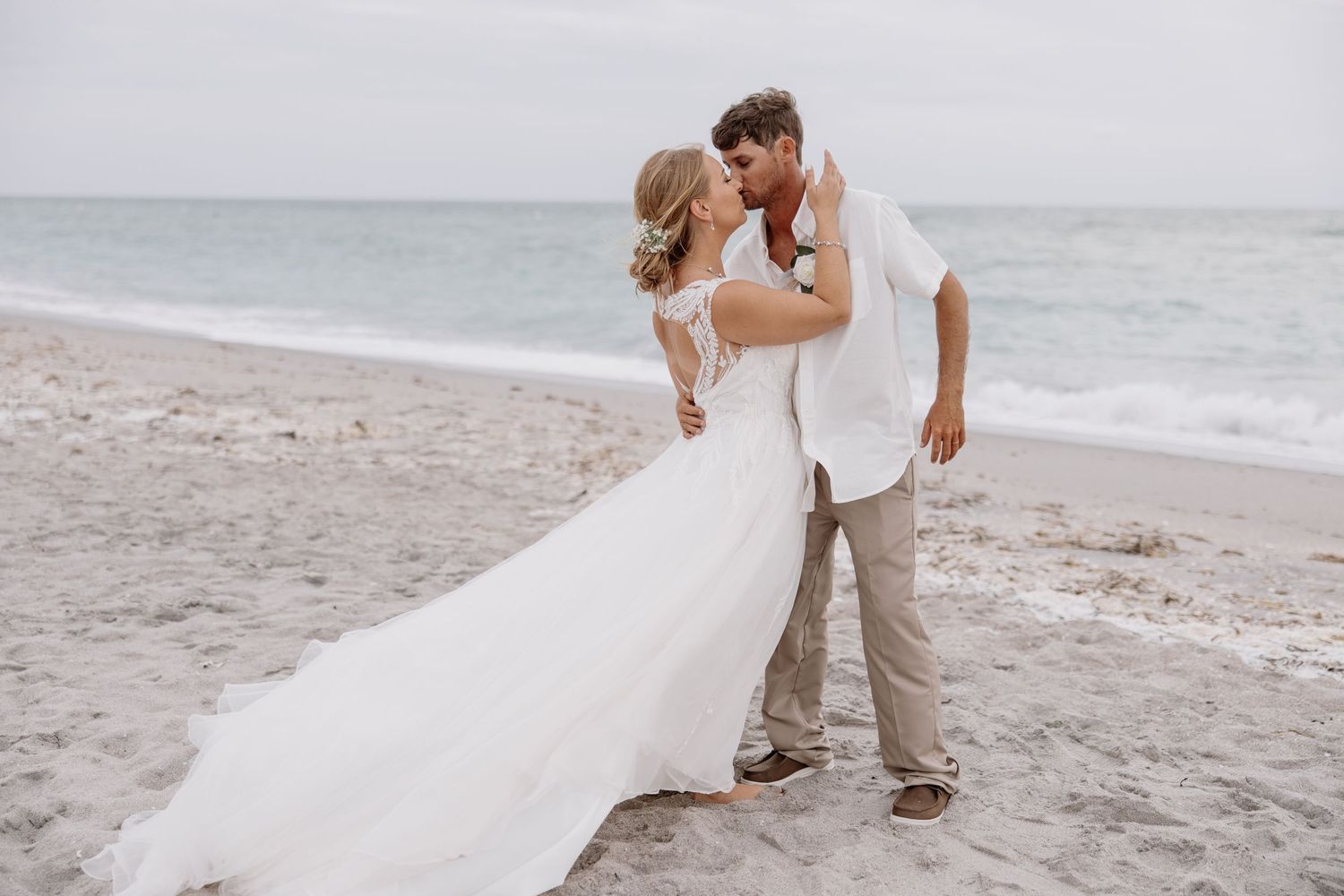 Wedding couple embracing on beach with flowing white dress catching the ocean breeze.