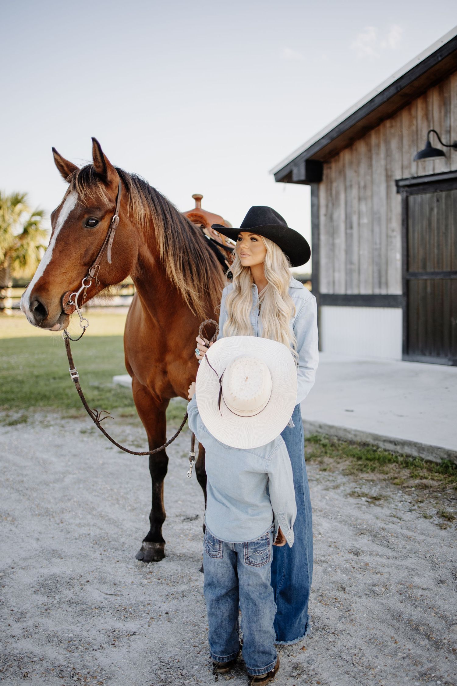 Woman in cowboy hat and denim stands with brown horse outside rustic barn.