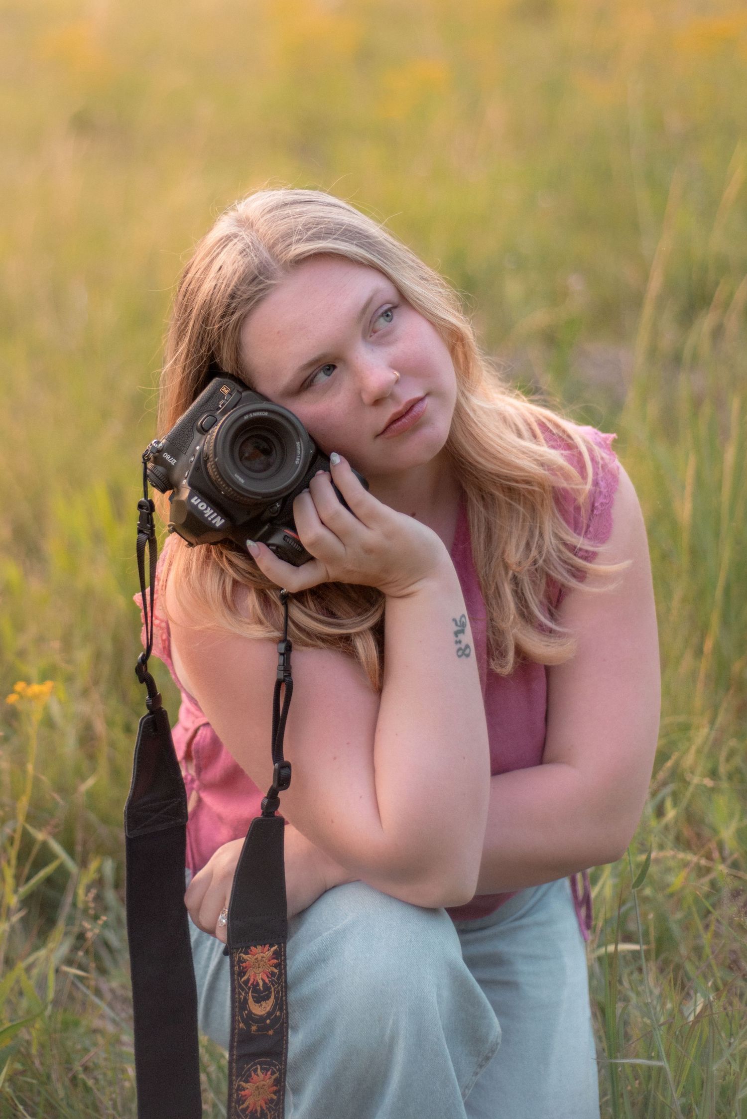 Young blonde woman holding a DSLR camera in a golden sunlit meadow, wearing a pink sleeveless top.