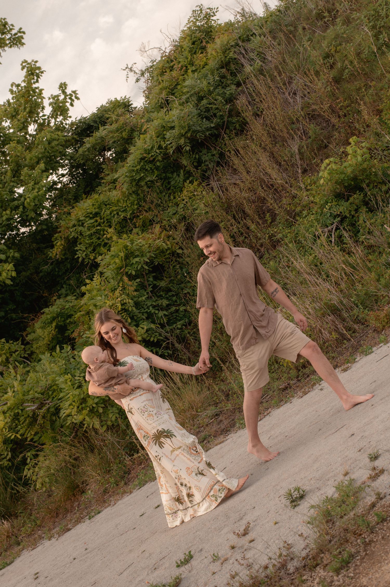 A young family walks hand-in-hand on a sandy path, mother holding a baby in a floral dress, father in tan shorts.