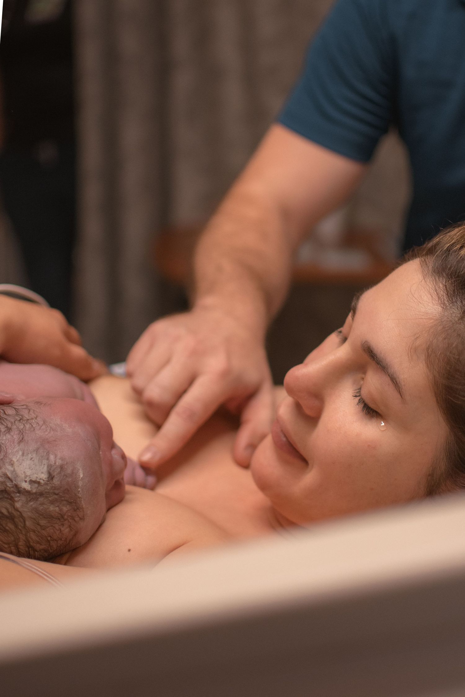 A mother holds her newborn baby skin-to-skin moments after birth, with a partner's hands visible offering support.