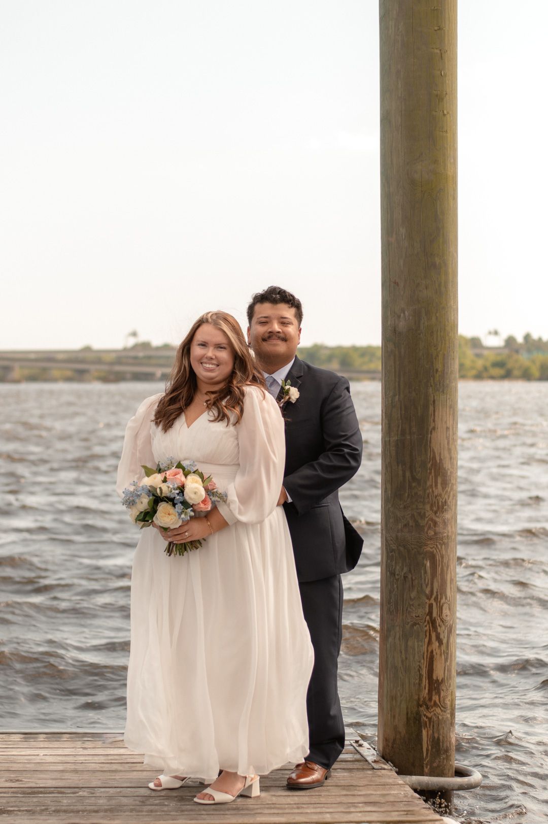 Couple in wedding attire posing on a waterfront dock, bride in white gown holding bouquet, groom in dark suit.
