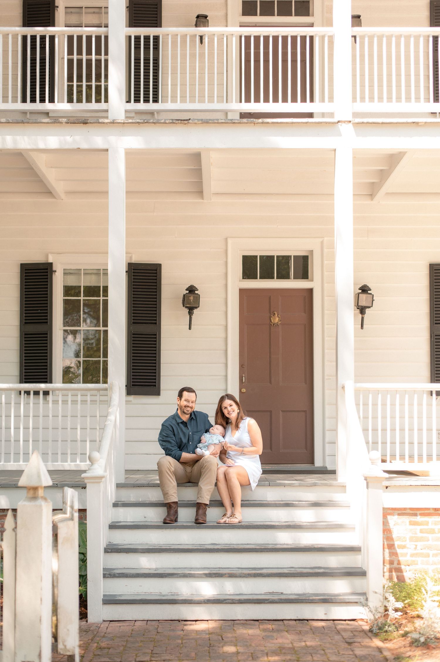 Couple sitting on steps of a white colonial home with black shutters, holding a newborn baby outdoors.