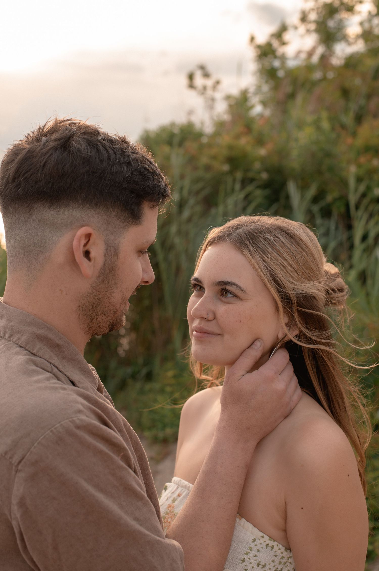 A couple shares a tender moment outdoors at golden hour, man gently holding womans face as she gazes up at him.