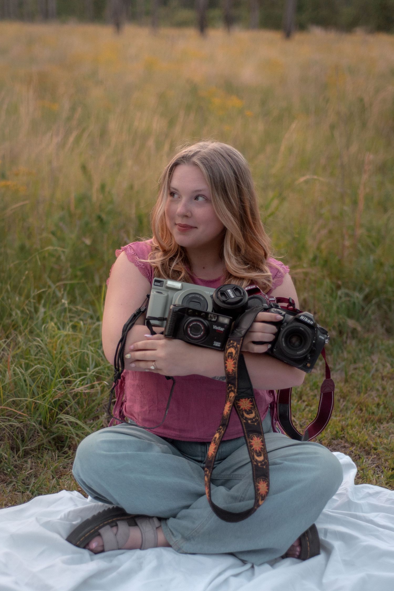 Young blonde woman sitting cross-legged in a meadow holding two cameras, wearing a pink top and gray jeans.