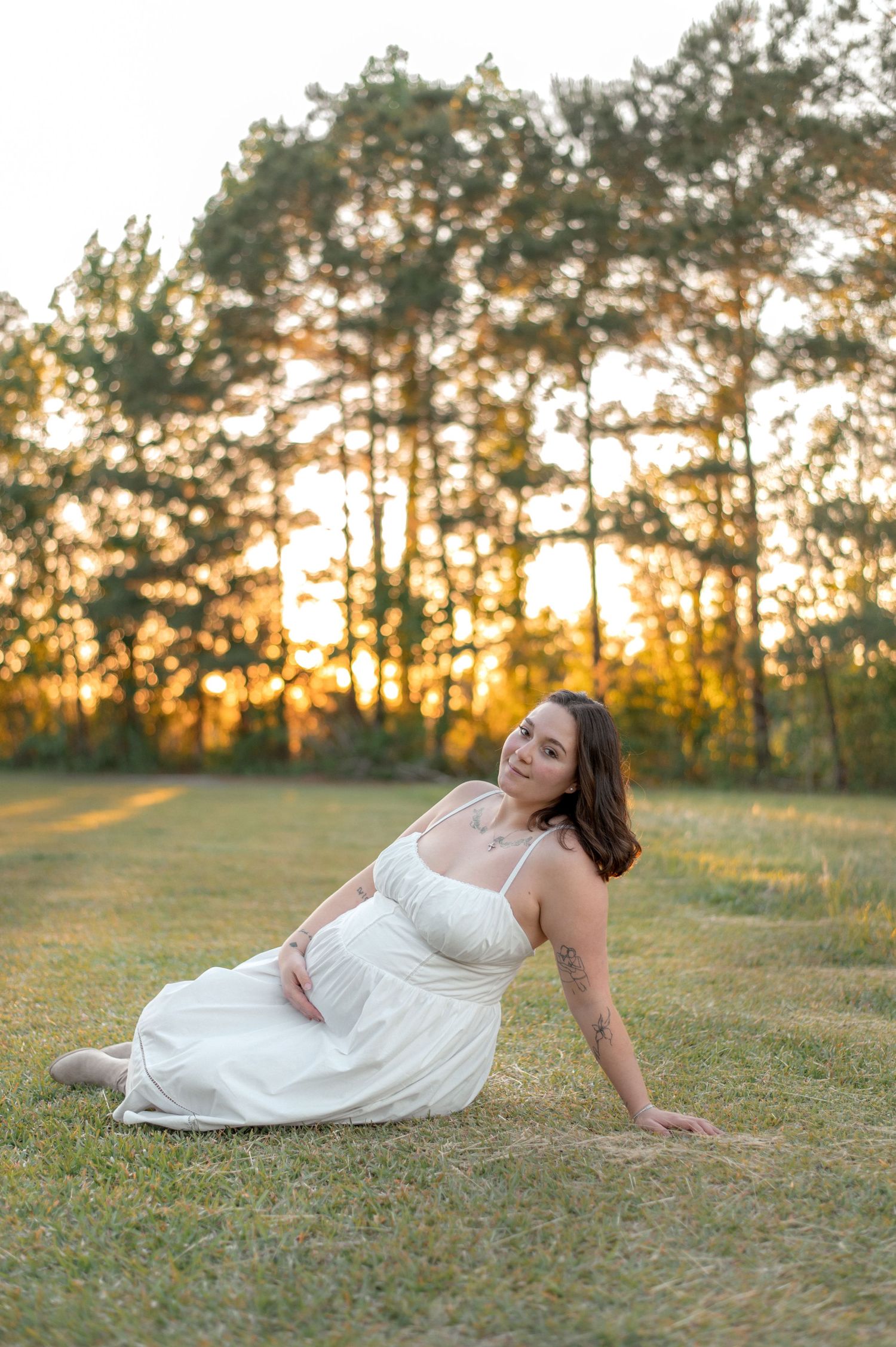 Woman in white dress sitting on grass at golden hour with tall pine trees glowing in sunset background.