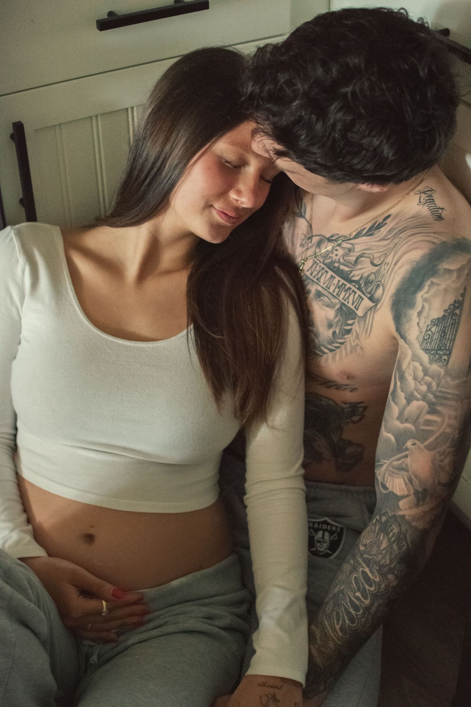 Pregnant woman in white crop top sitting as tattooed shirtless partner leans in tenderly in soft indoor light.