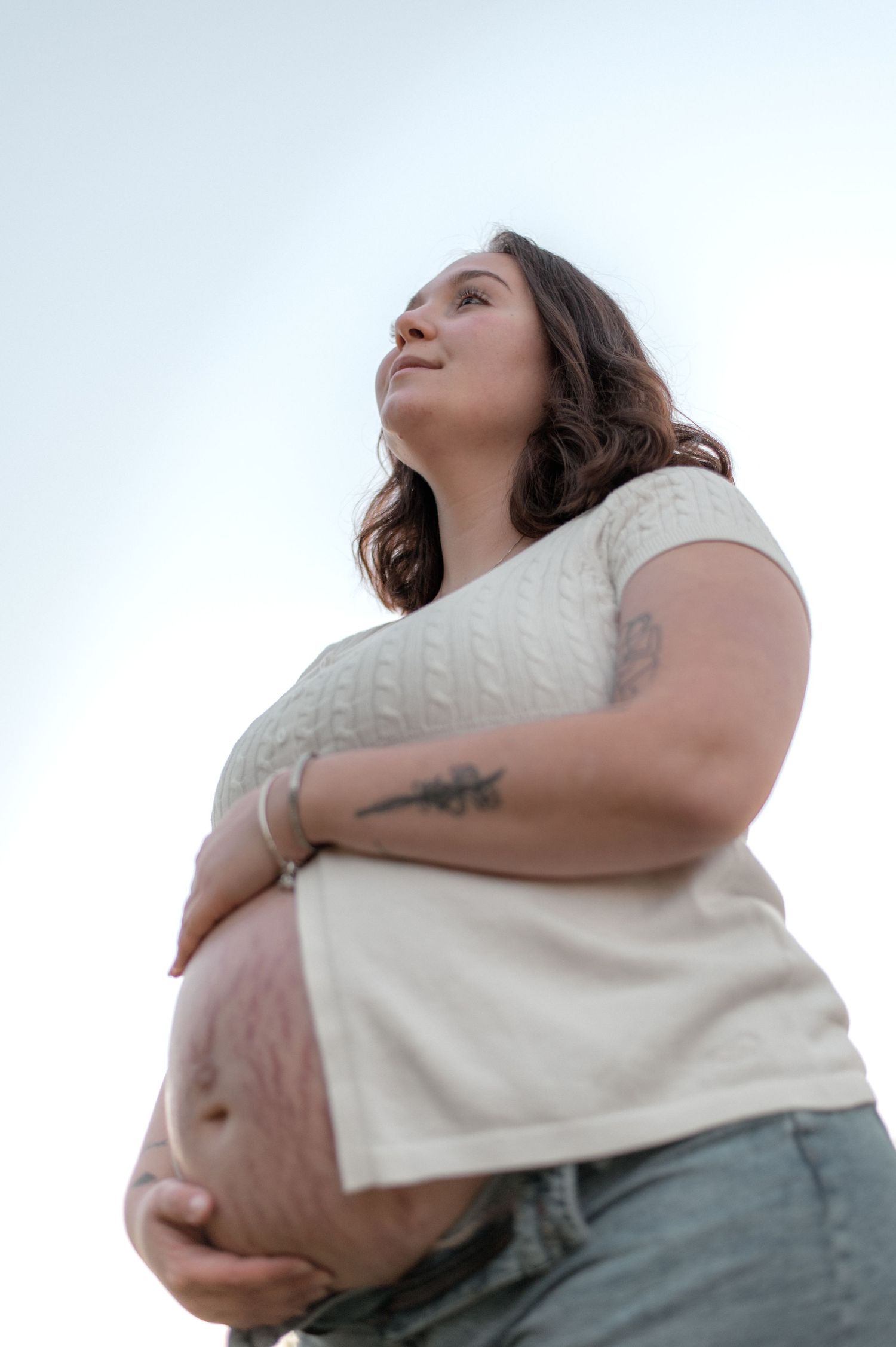 Pregnant woman in white top and jeans cradling her baby bump outdoors, shot from below against a bright sky.