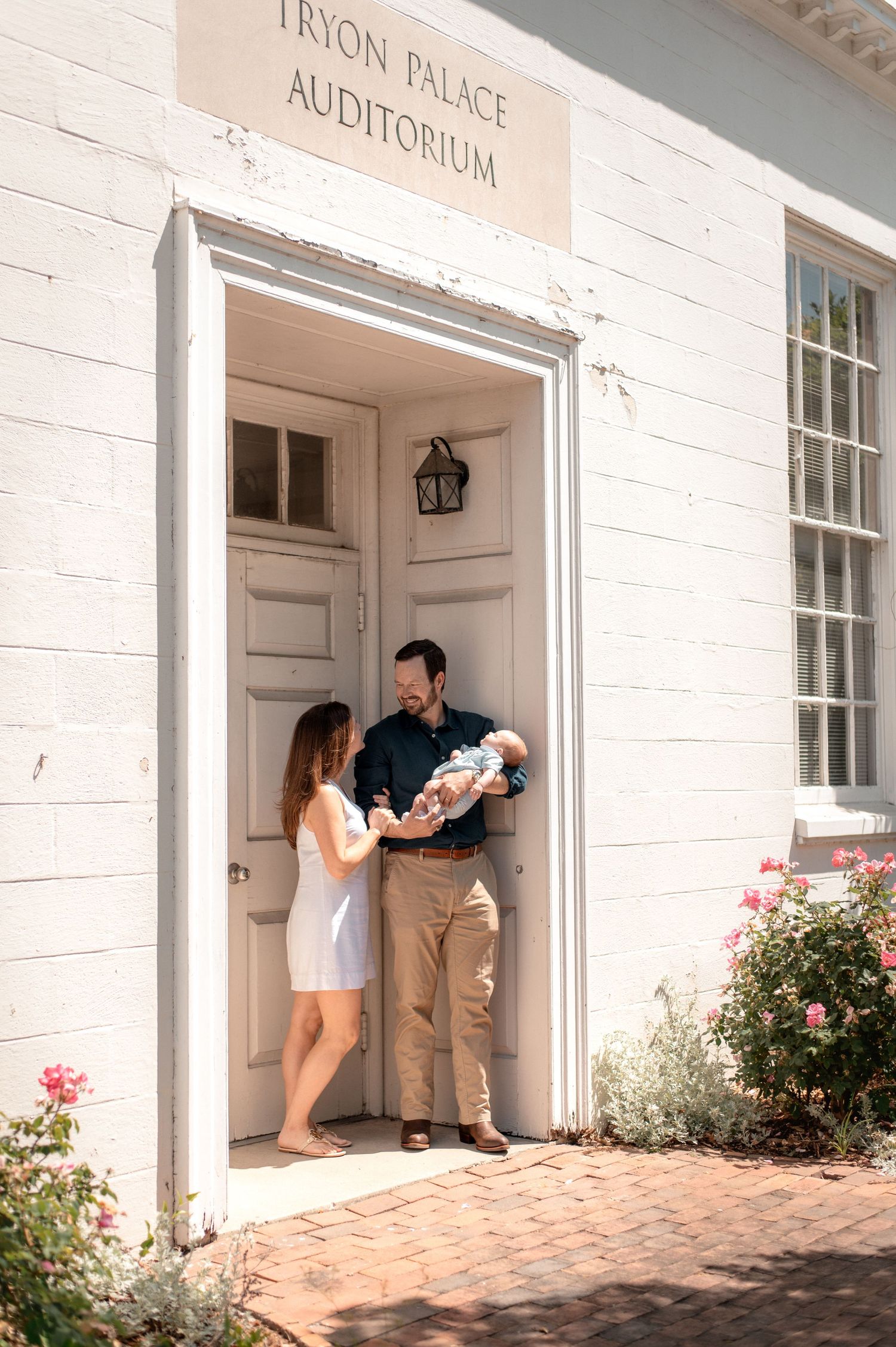 A couple stands in the doorway of Tryon Palace Auditorium holding a newborn baby, surrounded by blooming roses.