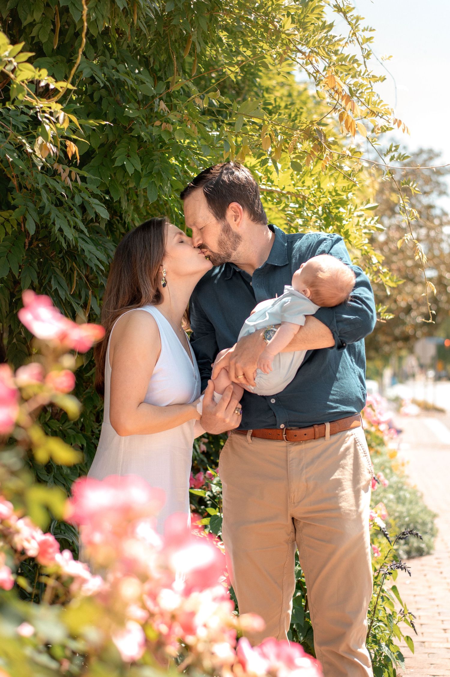A couple kisses outdoors while the man holds a newborn baby, surrounded by lush greenery and pink flowers.