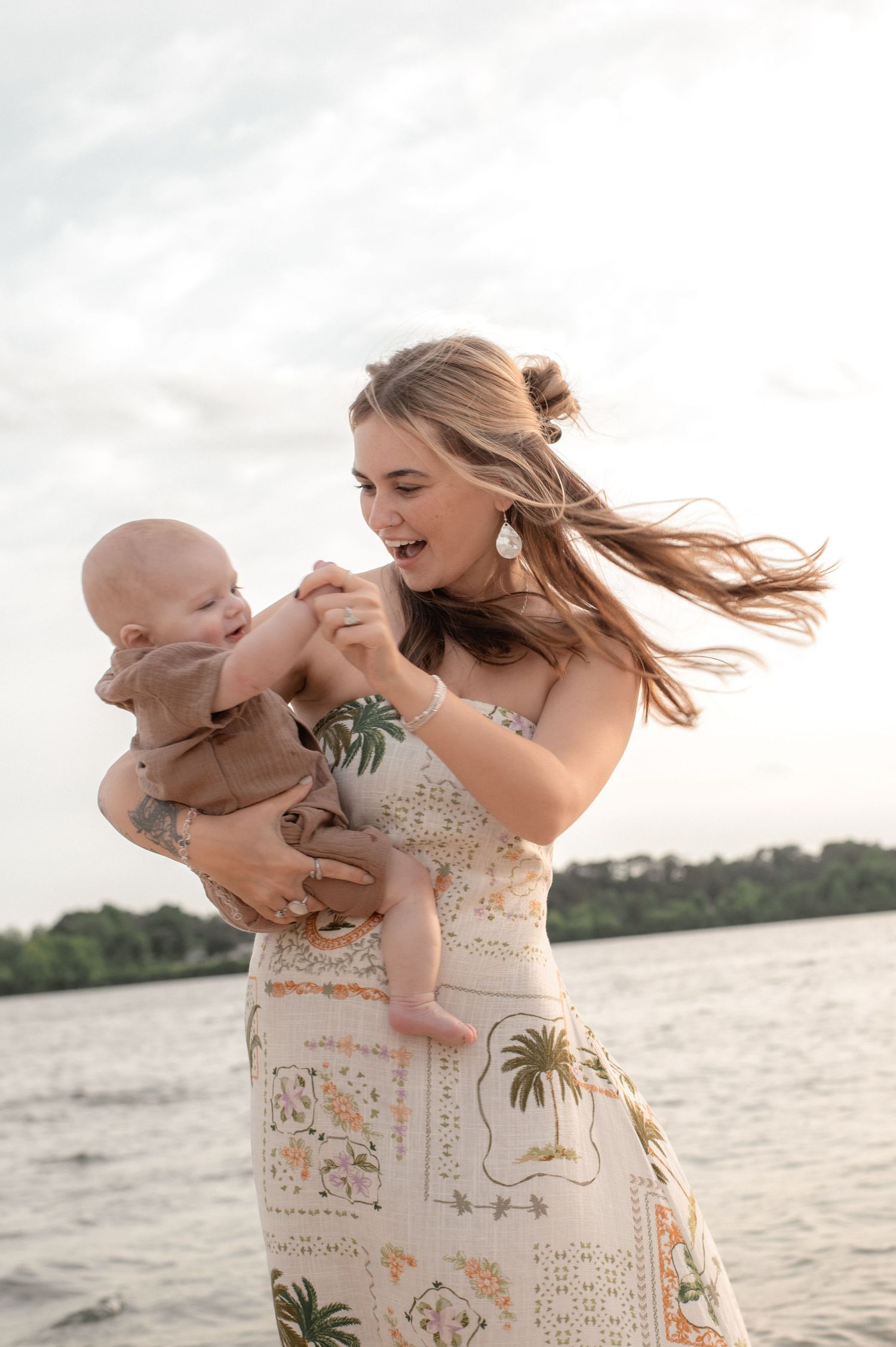 A joyful young mother in a floral dress holds her baby near a lake, laughing as her hair blows in the wind.