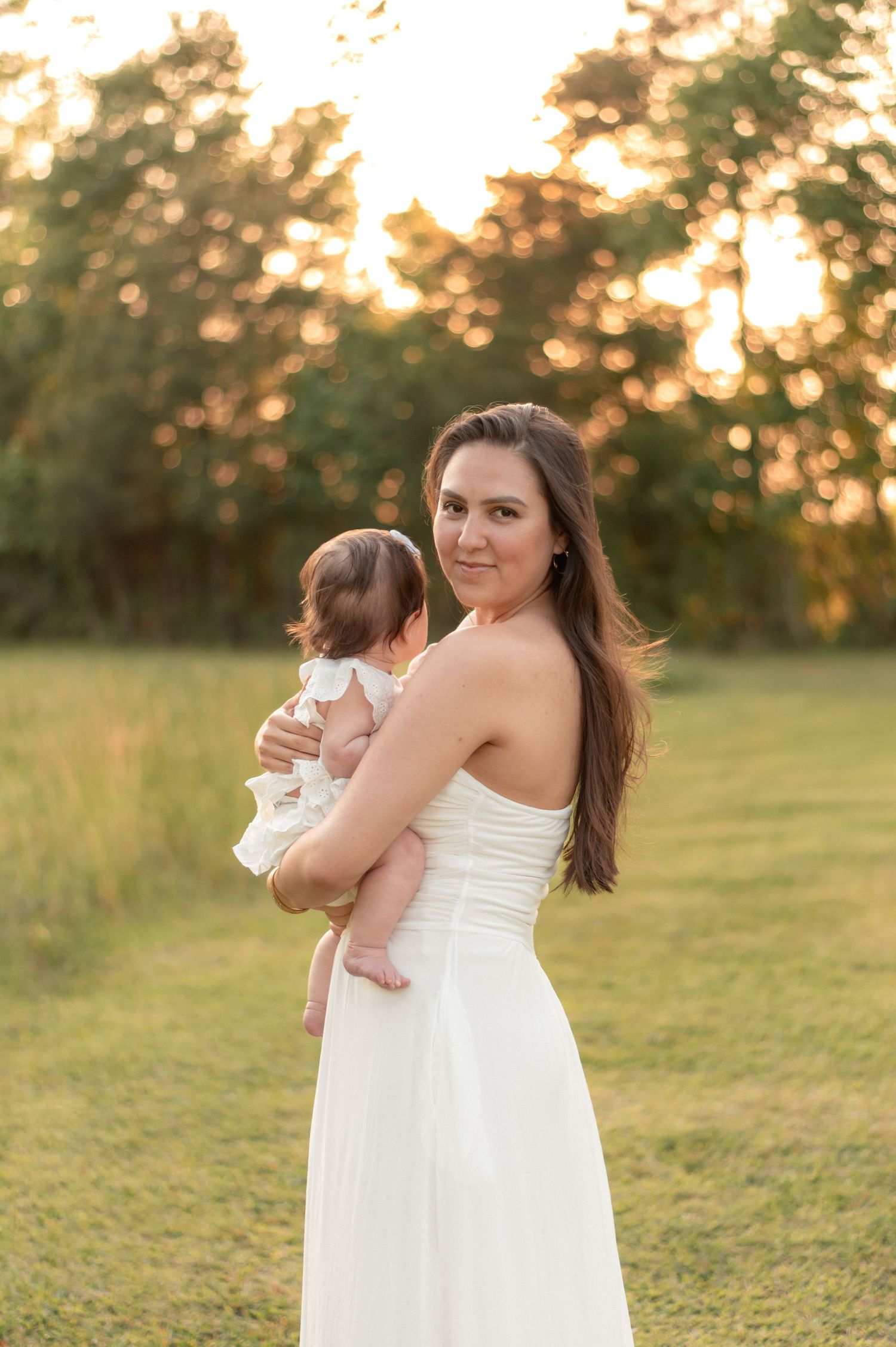 A woman in a white strapless gown holds her baby at golden hour in a lush green field.