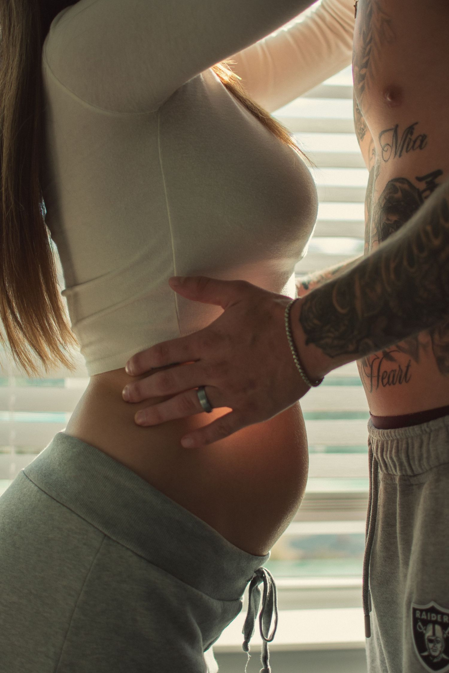 Close-up of pregnant woman's baby bump with tattooed partner's hand resting on her waist near sunlit window blinds.