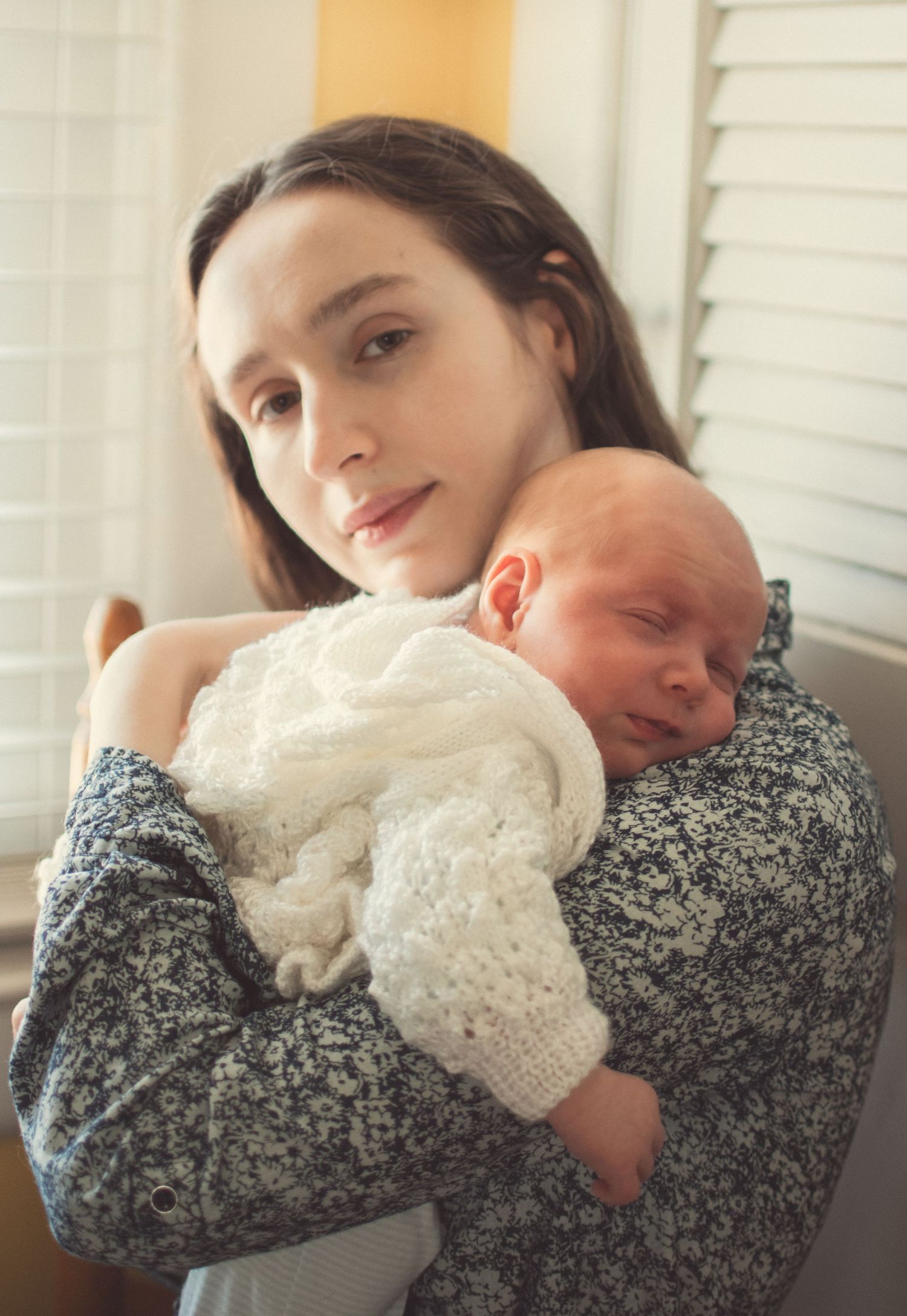 A serene new mother holds her peacefully sleeping newborn wrapped in a white knit blanket.