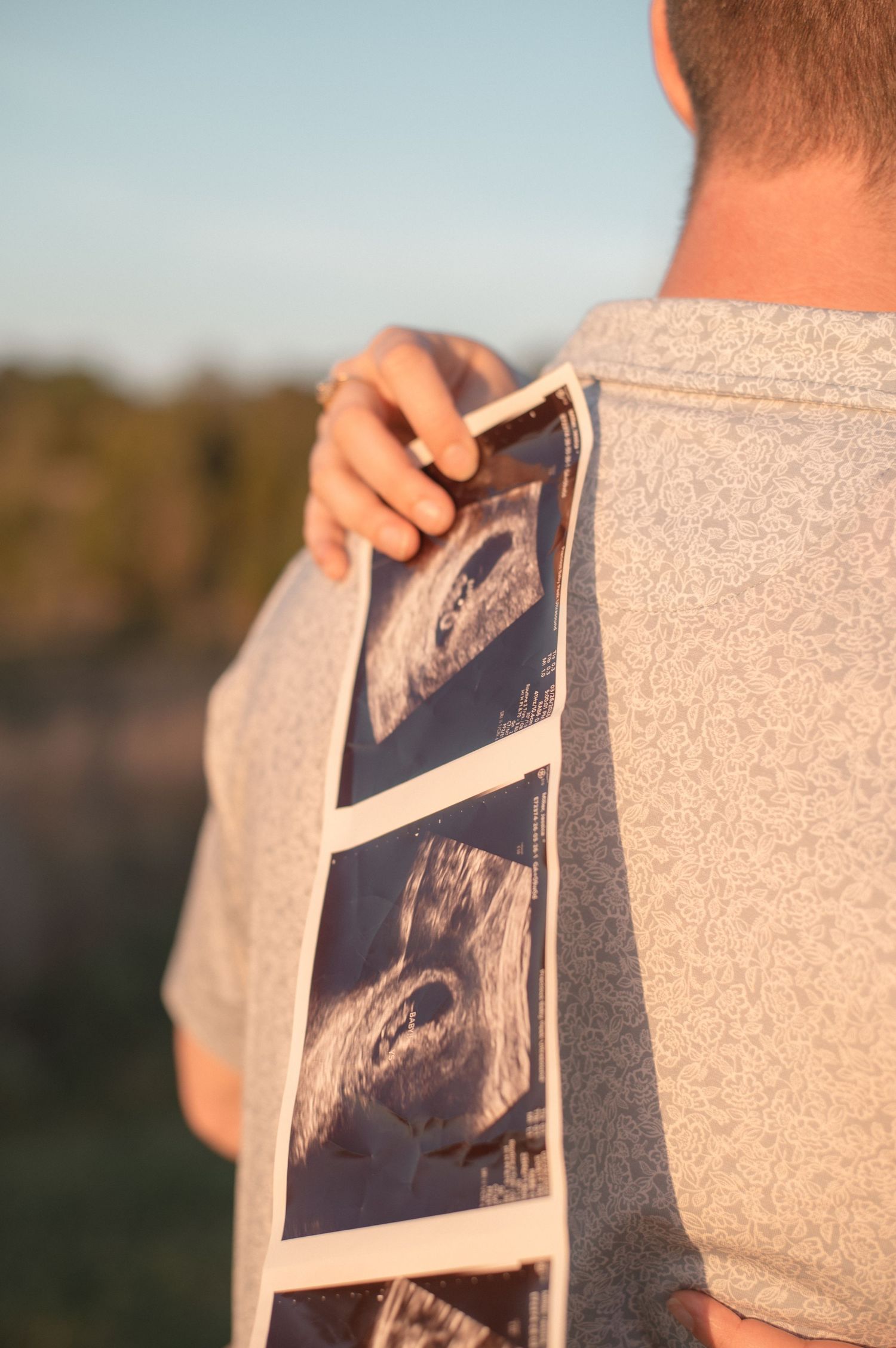 Man holding ultrasound photos over his shoulder outdoors at golden hour, announcing pregnancy.