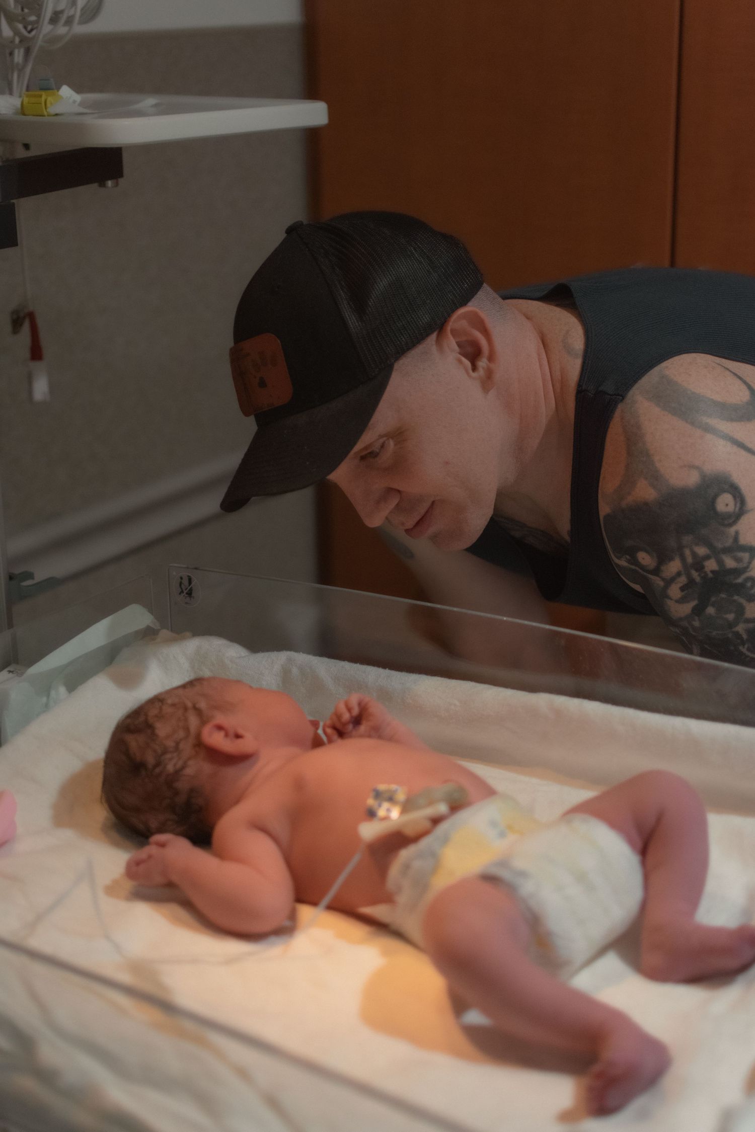 A tattooed father in a baseball cap gazes lovingly at his newborn baby lying in a hospital bassinet.