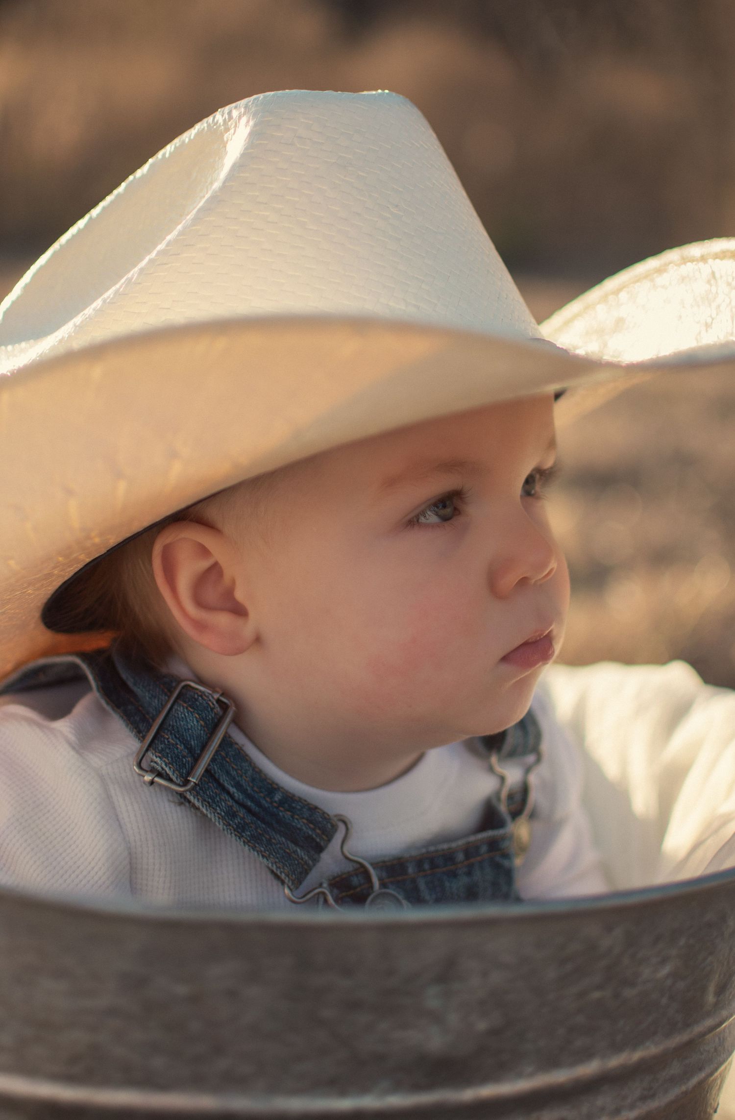Close-up portrait of a cute baby wearing a white cowboy hat and denim overalls in warm golden light.