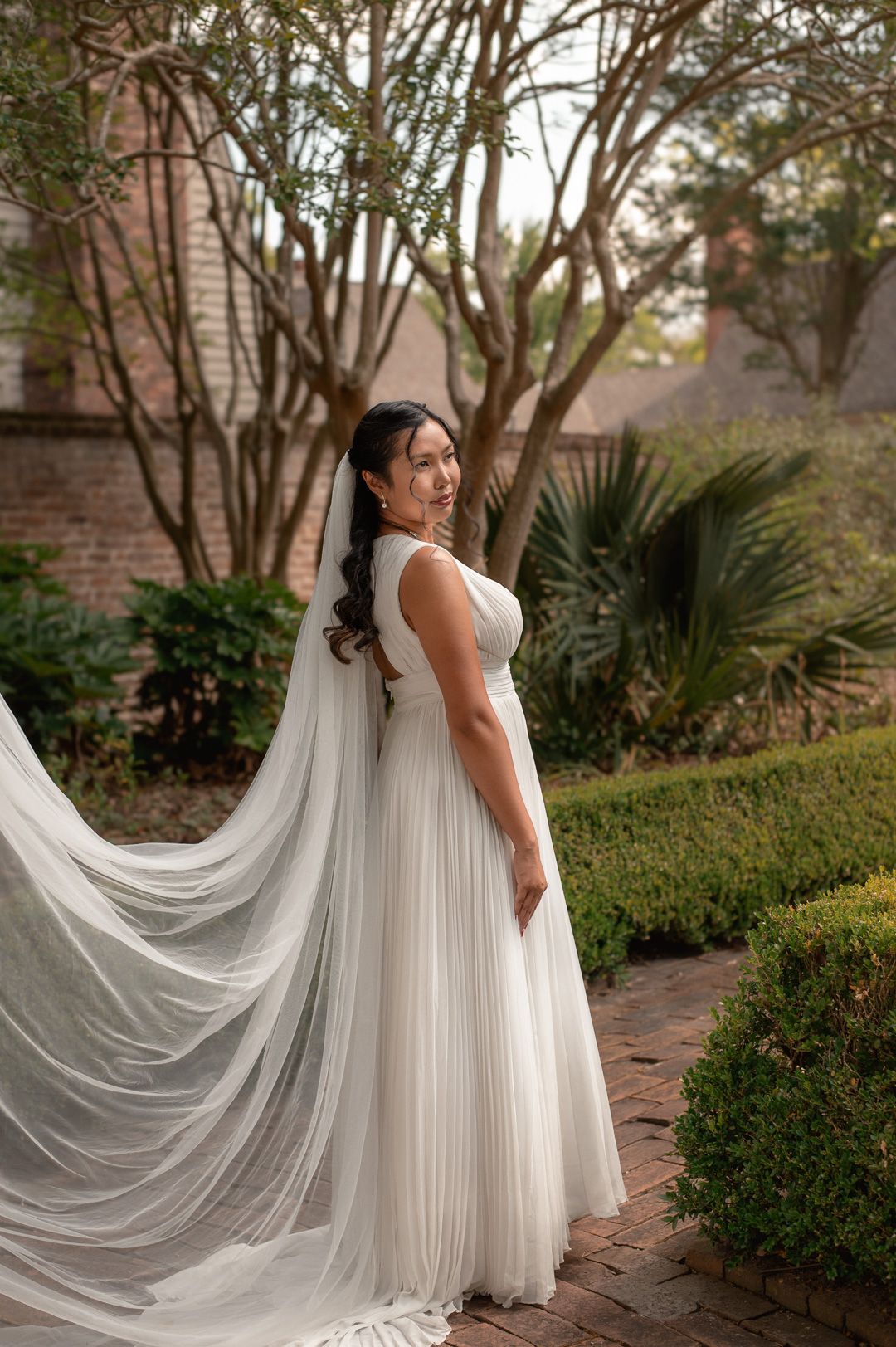 Bride in a flowing white pleated gown with a long veil, smiling over her shoulder in a lush garden setting.