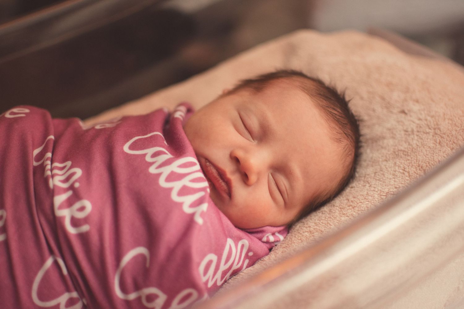 Peaceful newborn baby sleeping in hospital bassinet wrapped in pink personalized swaddle blanket.