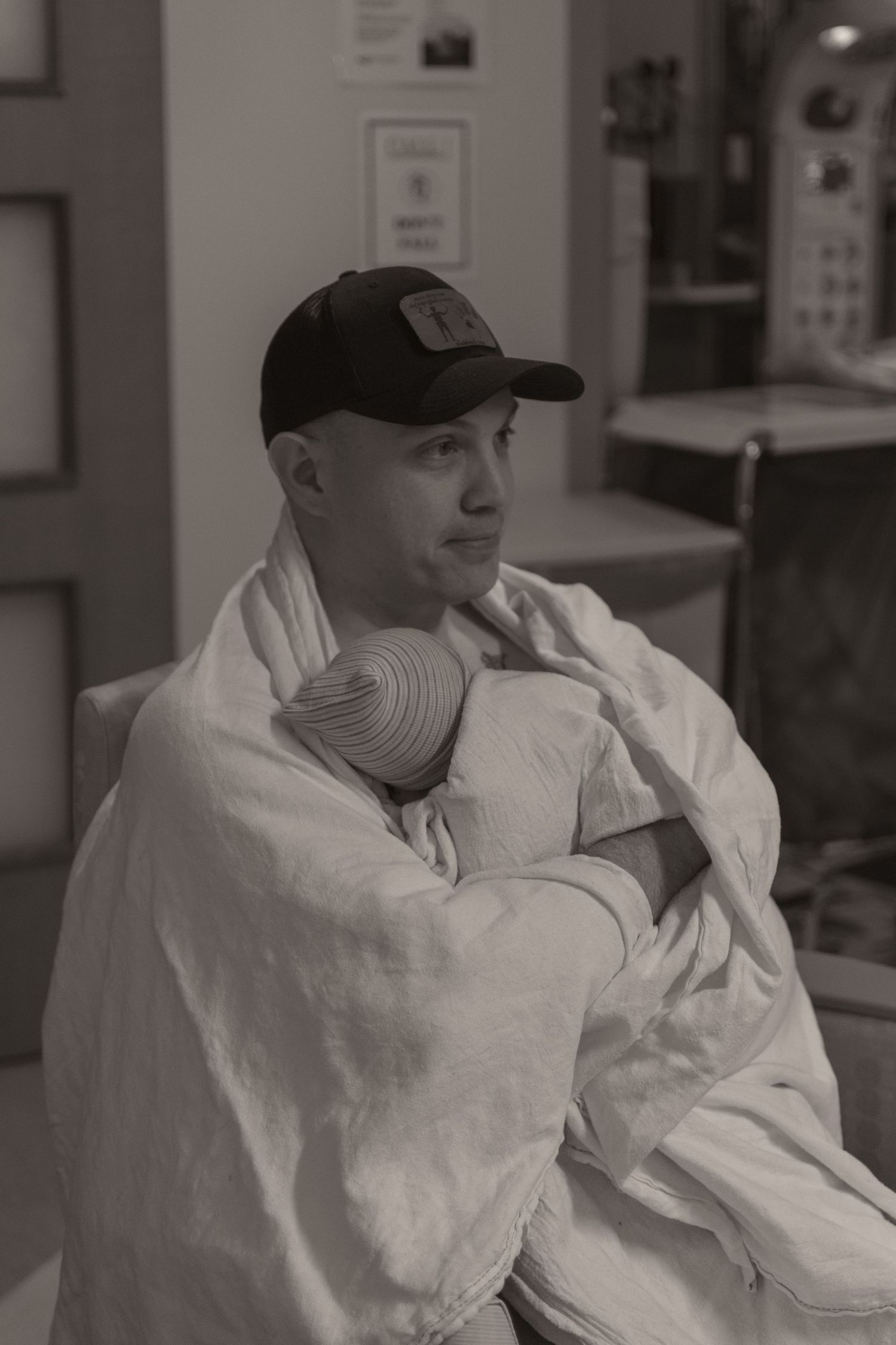 Black and white photo of a man in a baseball cap holding a newborn baby wrapped in a white blanket in a hospital room.