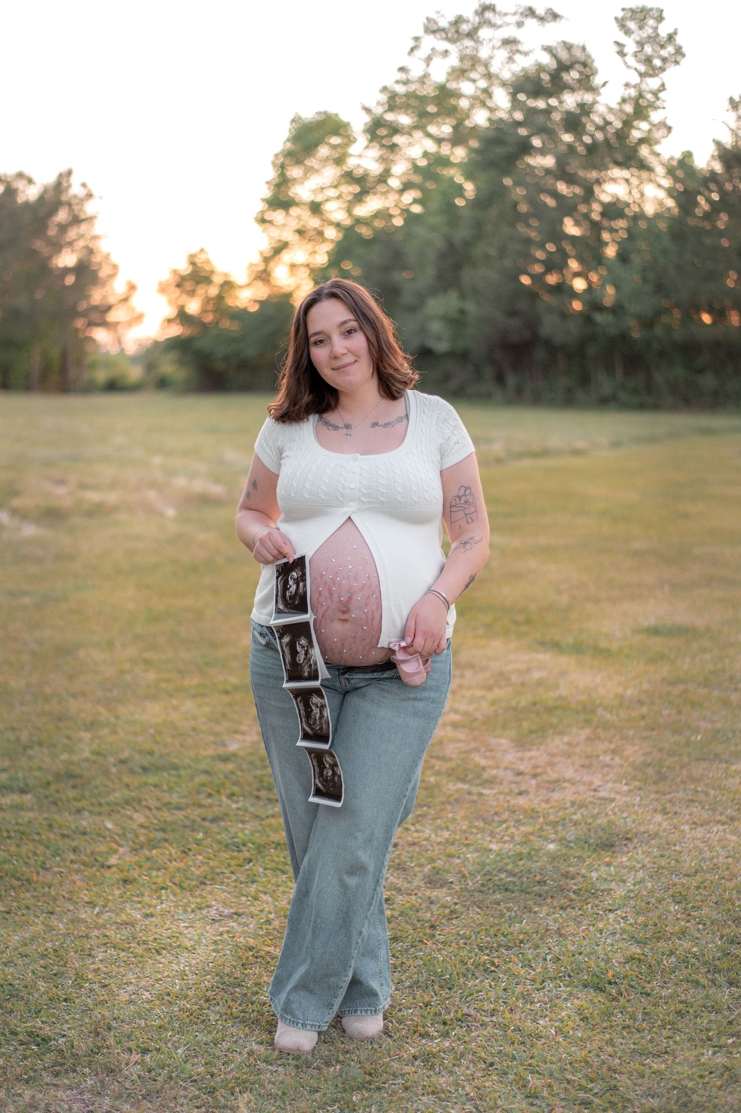 Pregnant woman in white top and jeans holding ultrasound photos outdoors at golden hour in a grassy field.