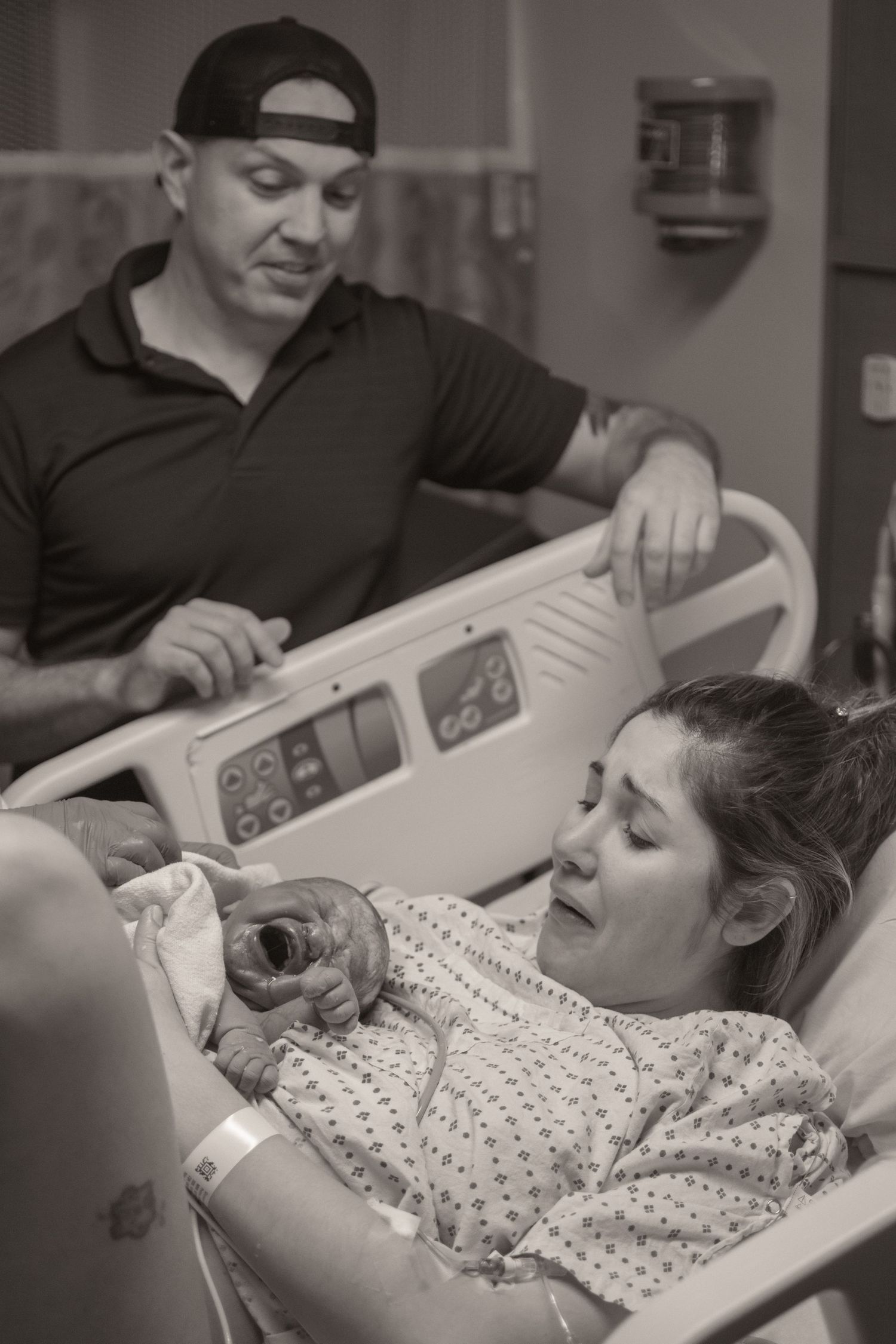 Black and white photo of a mother in hospital bed holding her newborn baby with father standing beside her.