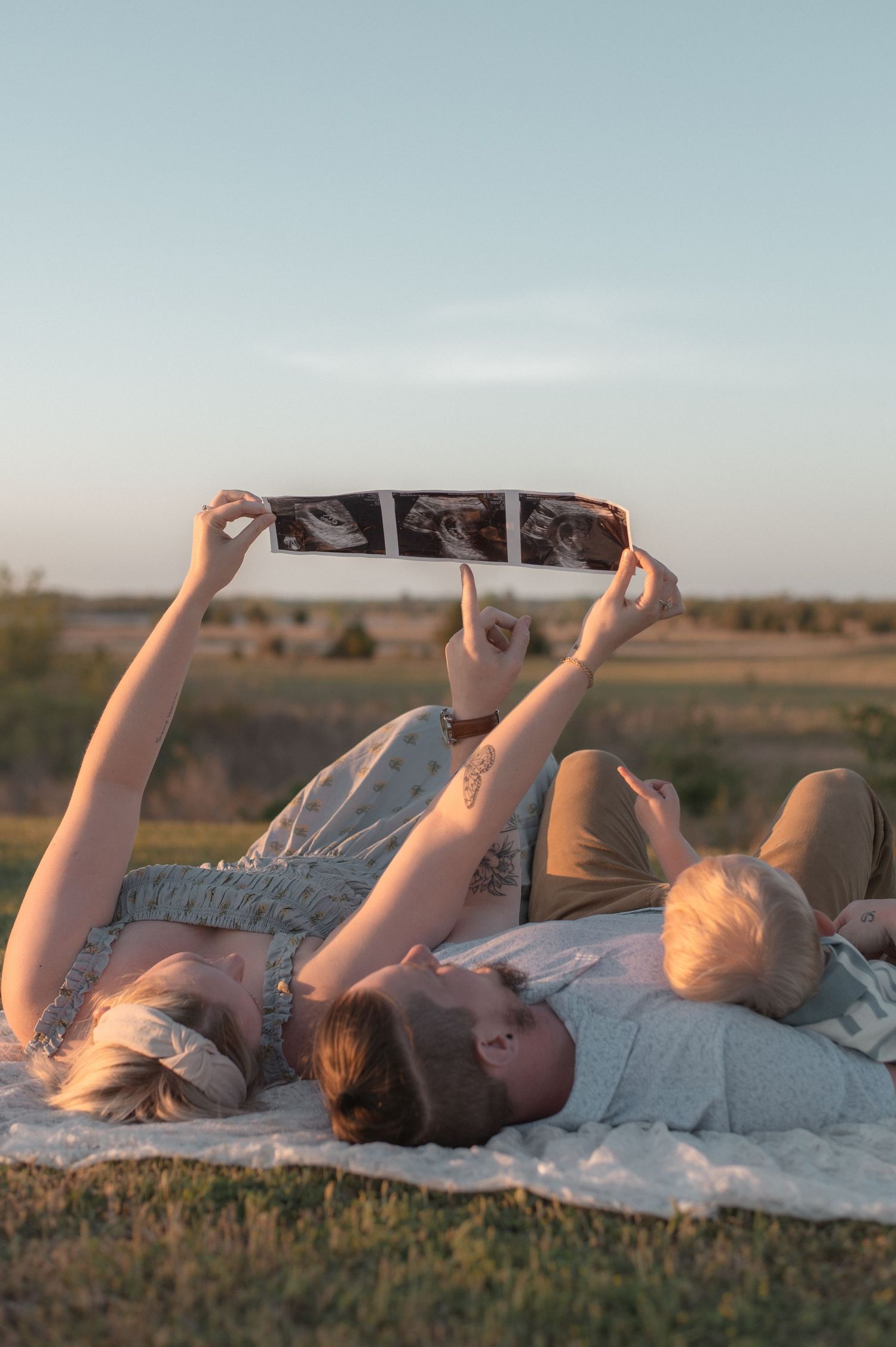 Family lying on blanket outdoors holding ultrasound photos up in golden hour light during pregnancy announcement.