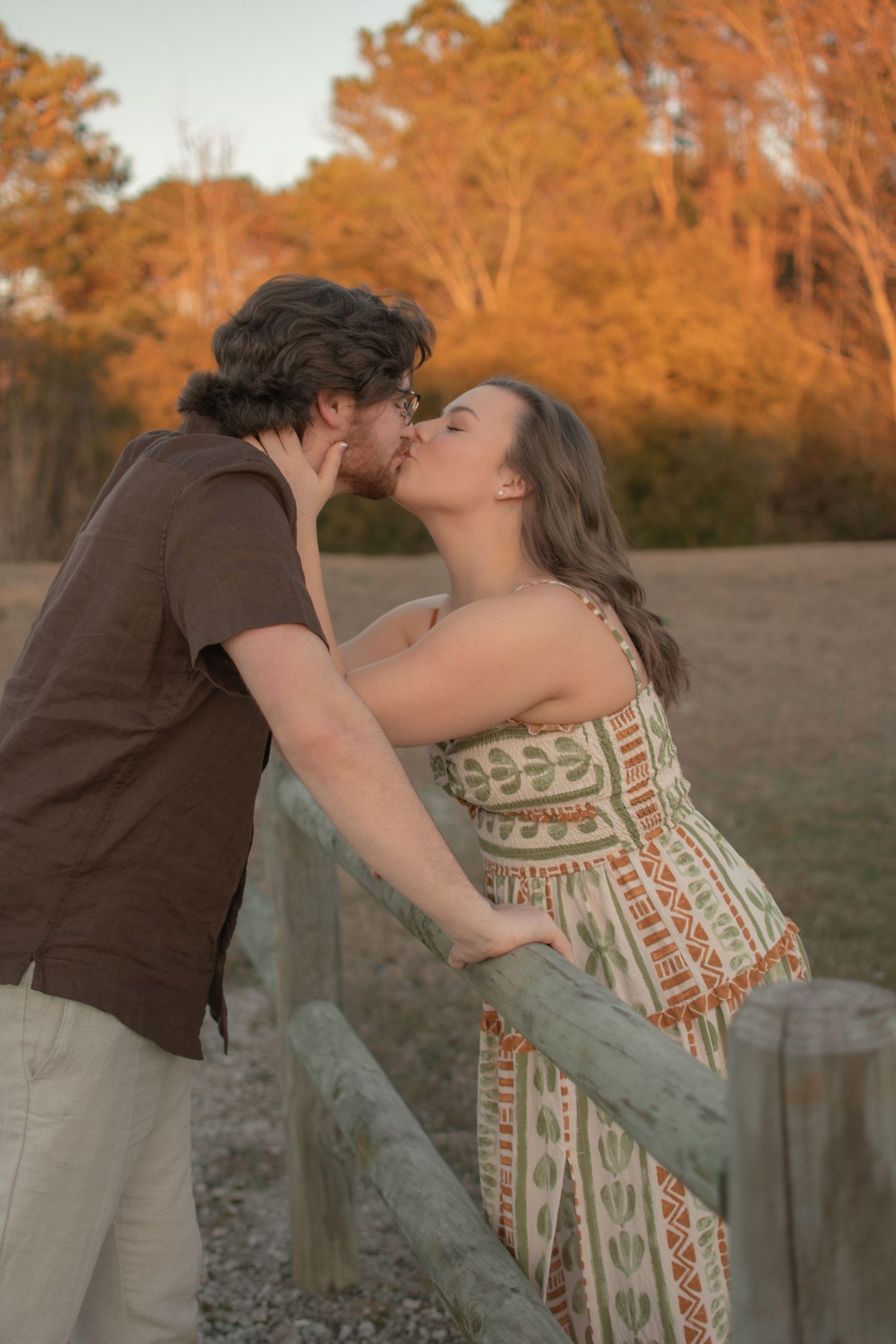 A couple shares a romantic kiss leaning against a wooden fence during a warm autumn sunset.