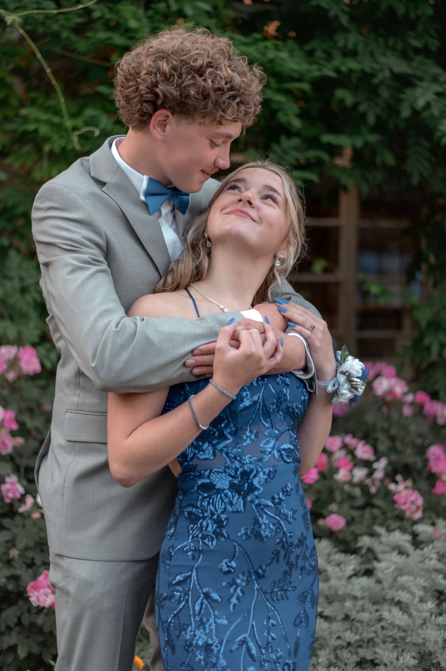 Teen couple in prom attire, boy in gray suit with blue bow tie embracing girl in blue floral dress outdoors.